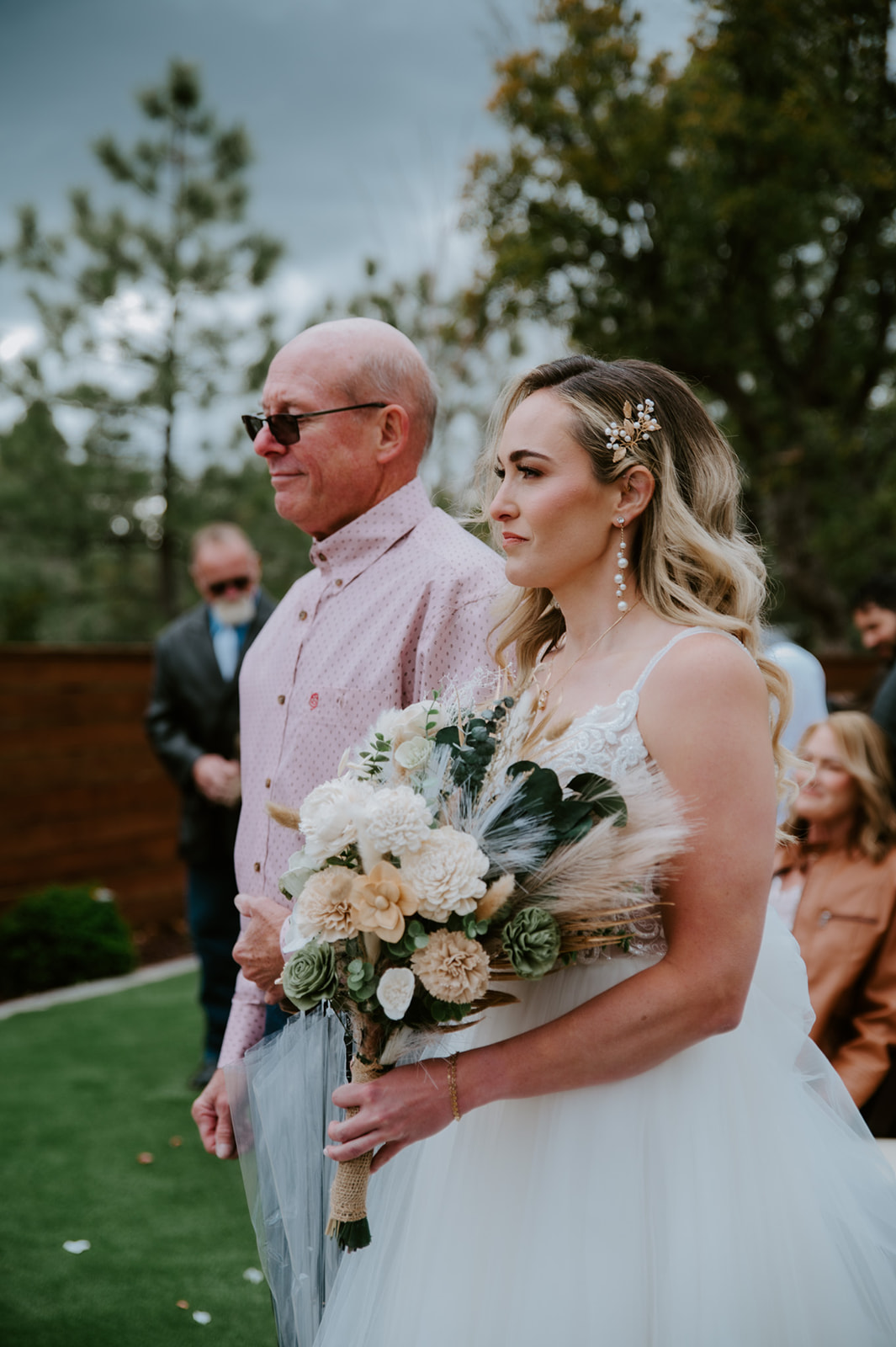 Bride holding a neutral-toned bouquet walks down the aisle before the outdoor wedding ceremony.
