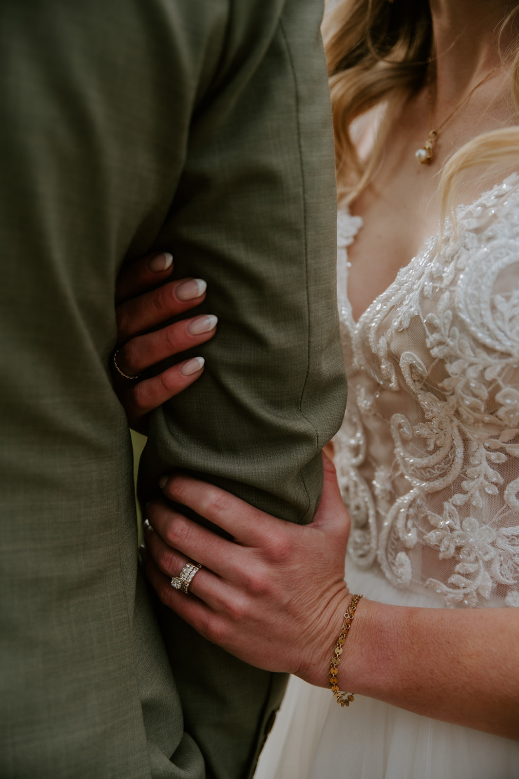 Close-up of the bride holding the groom's arm, highlighting her wedding ring and lace dress details.

