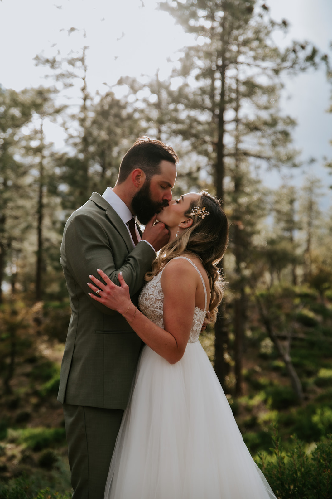 Bride and groom share a kiss beneath warm sunlight filtering through the trees.
