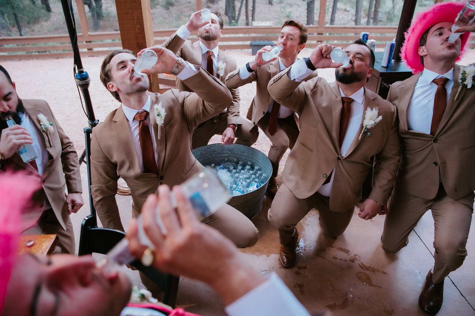 Groomsmen kneel together while taking celebratory shots during the wedding reception, with bright pink cowboy hats adding a fun touch.
