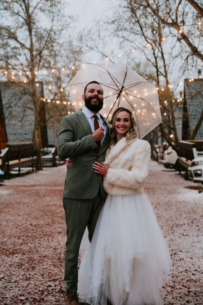 Bride and groom posing beneath café lights with a clear umbrella outside their cabin after the ceremony.