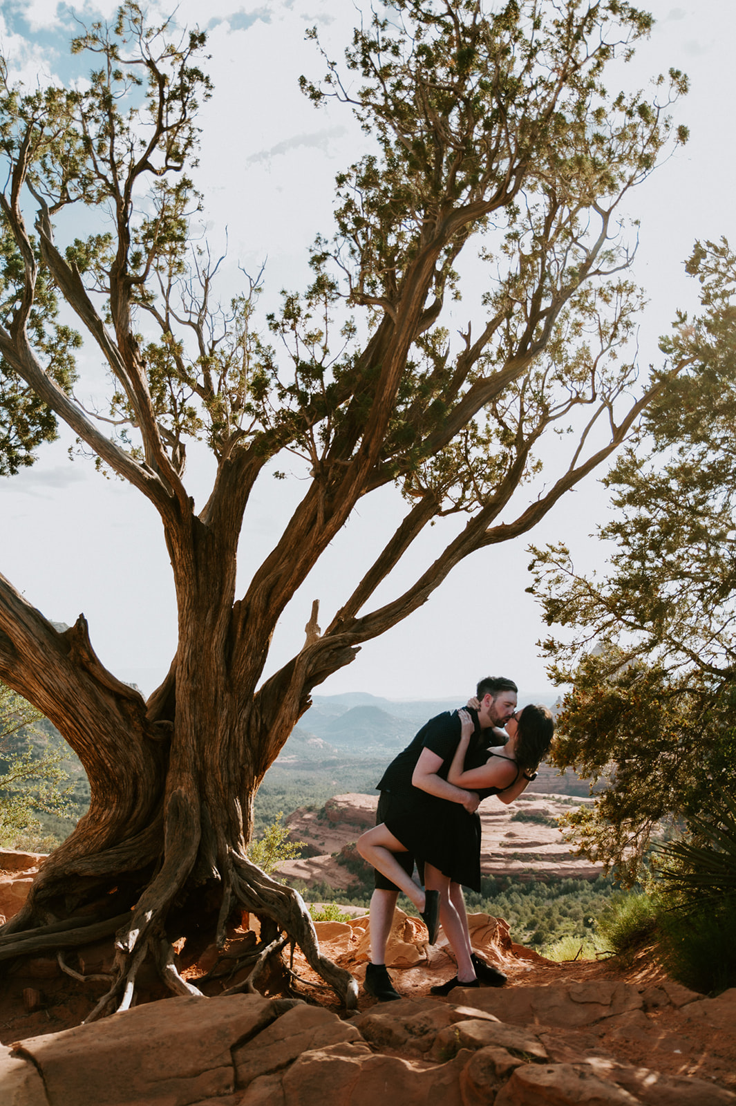 Couple sharing a kiss beneath a twisted juniper tree overlooking Sedona's desert landscape.