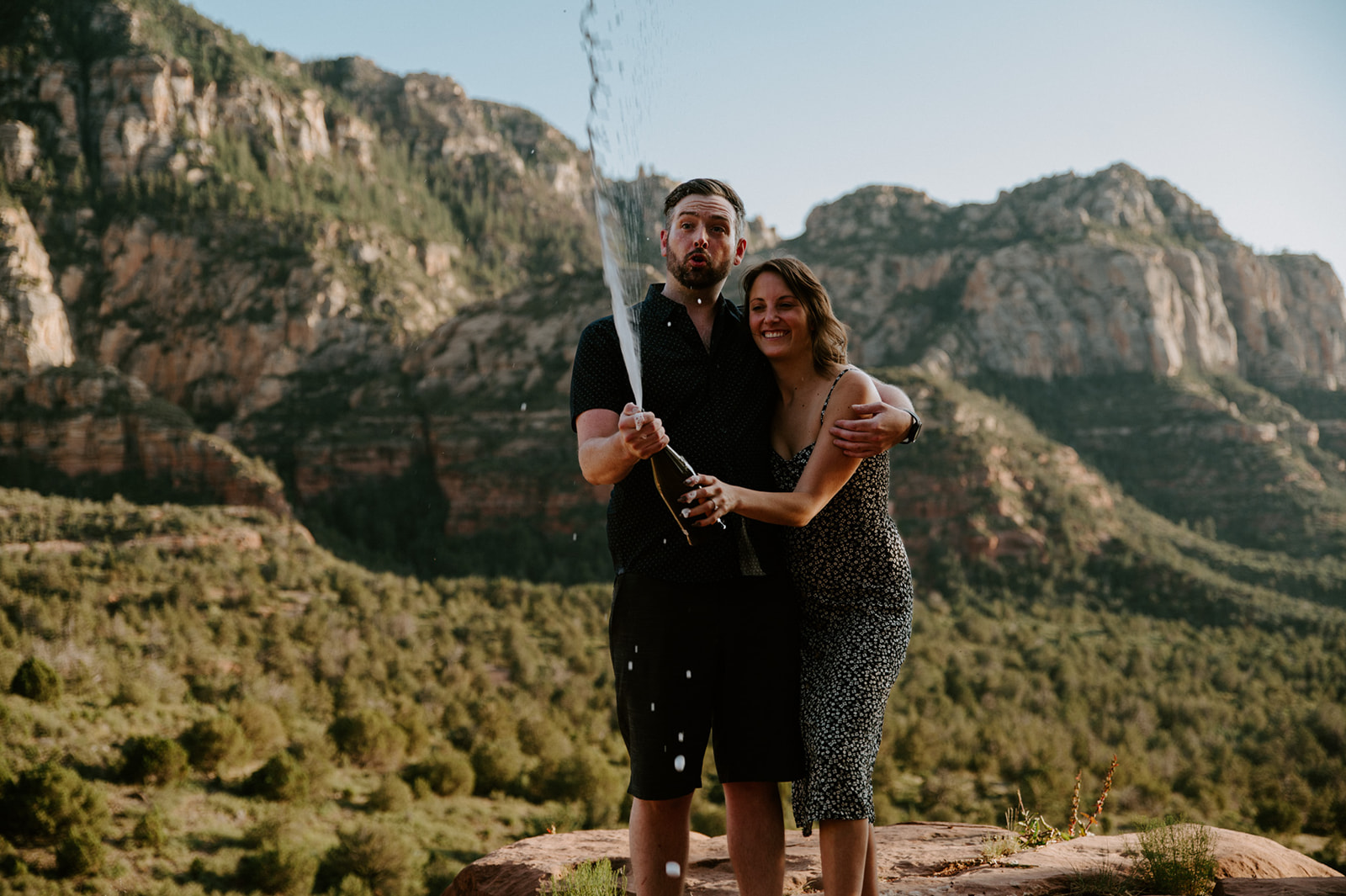 Couple celebrating with champagne after their Merry Go Round Rock Sedona engagement session overlooking the cliffs of Sedona.