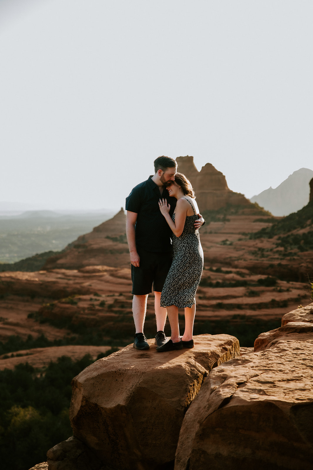 Couple embracing on a sandstone overlook at Merry Go Round Rock Sedona with iconic red rock formations glowing in the evening light.