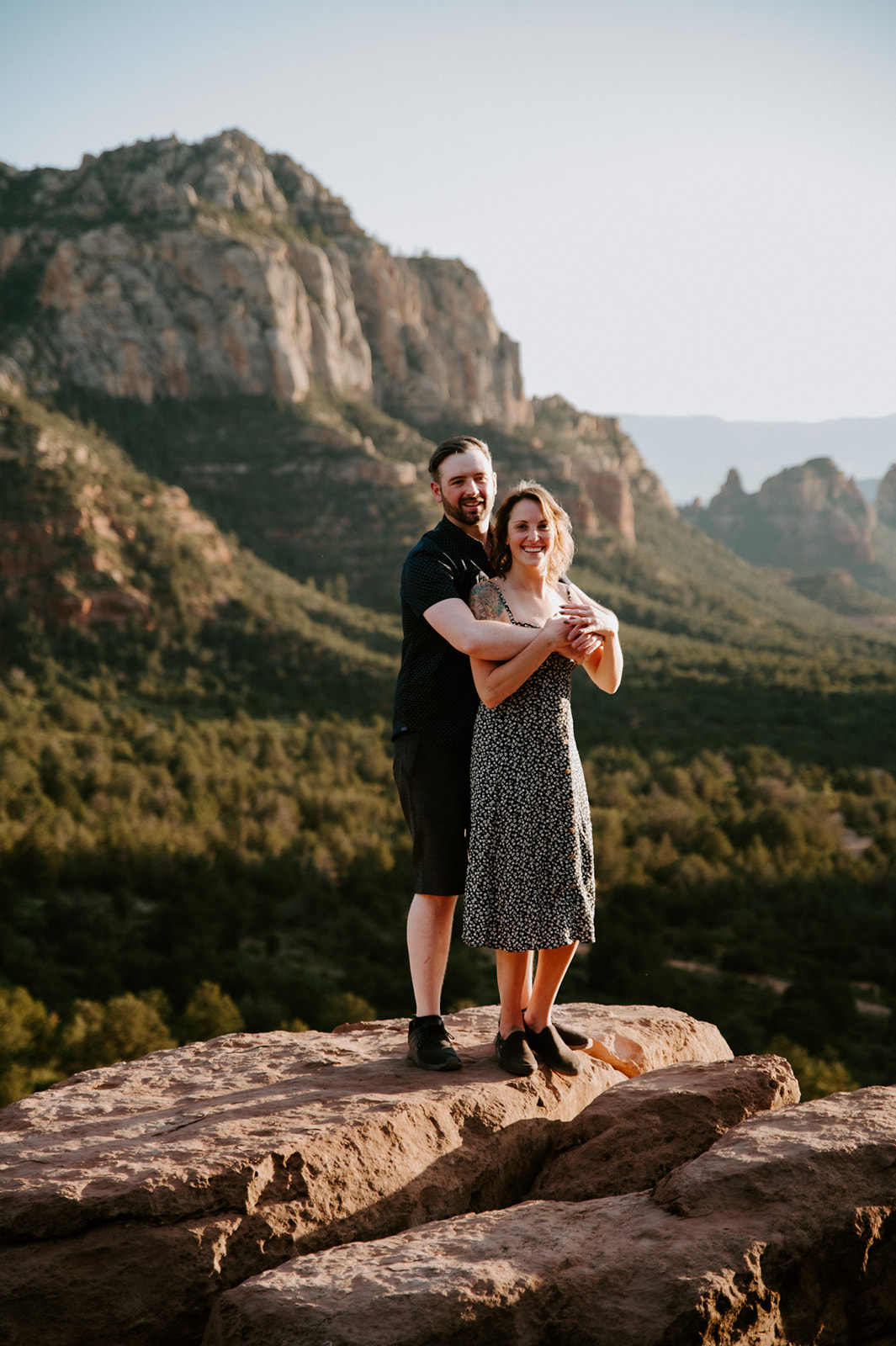 Couple embracing on a sandstone overlook with towering red rock cliffs surrounding Merry Go Round Rock Sedona.