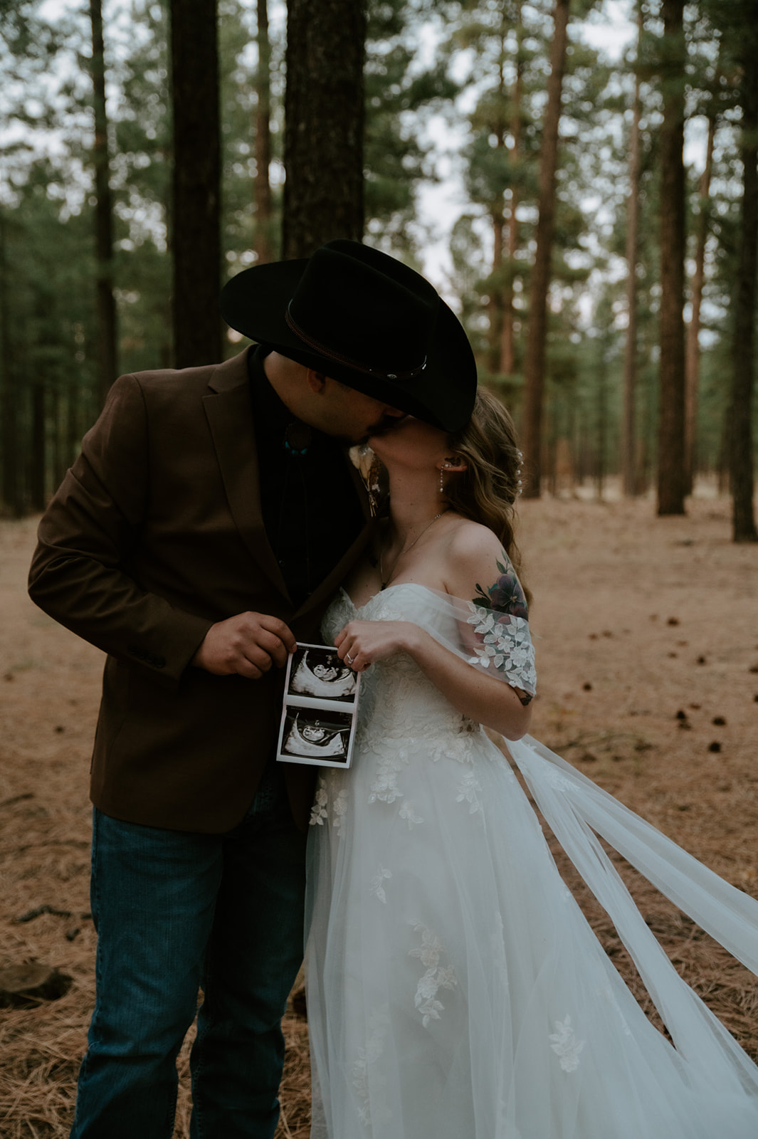 Bride and groom announcing their pregnancy during wedding portraits in the Arizona pine forest while holding ultrasound photos.