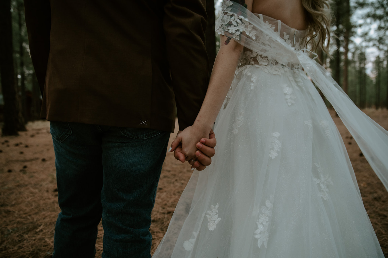 Close-up of newlyweds holding hands as the bride's lace wedding dress and veil flow through a peaceful Arizona forest.