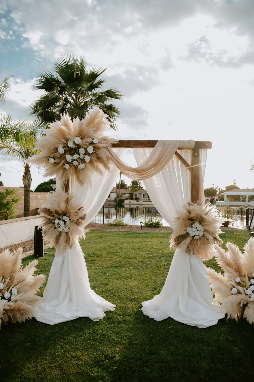 Outdoor wedding arch draped in ivory fabric and decorated with pampas grass and white flowers for backyard wedding ideas.
