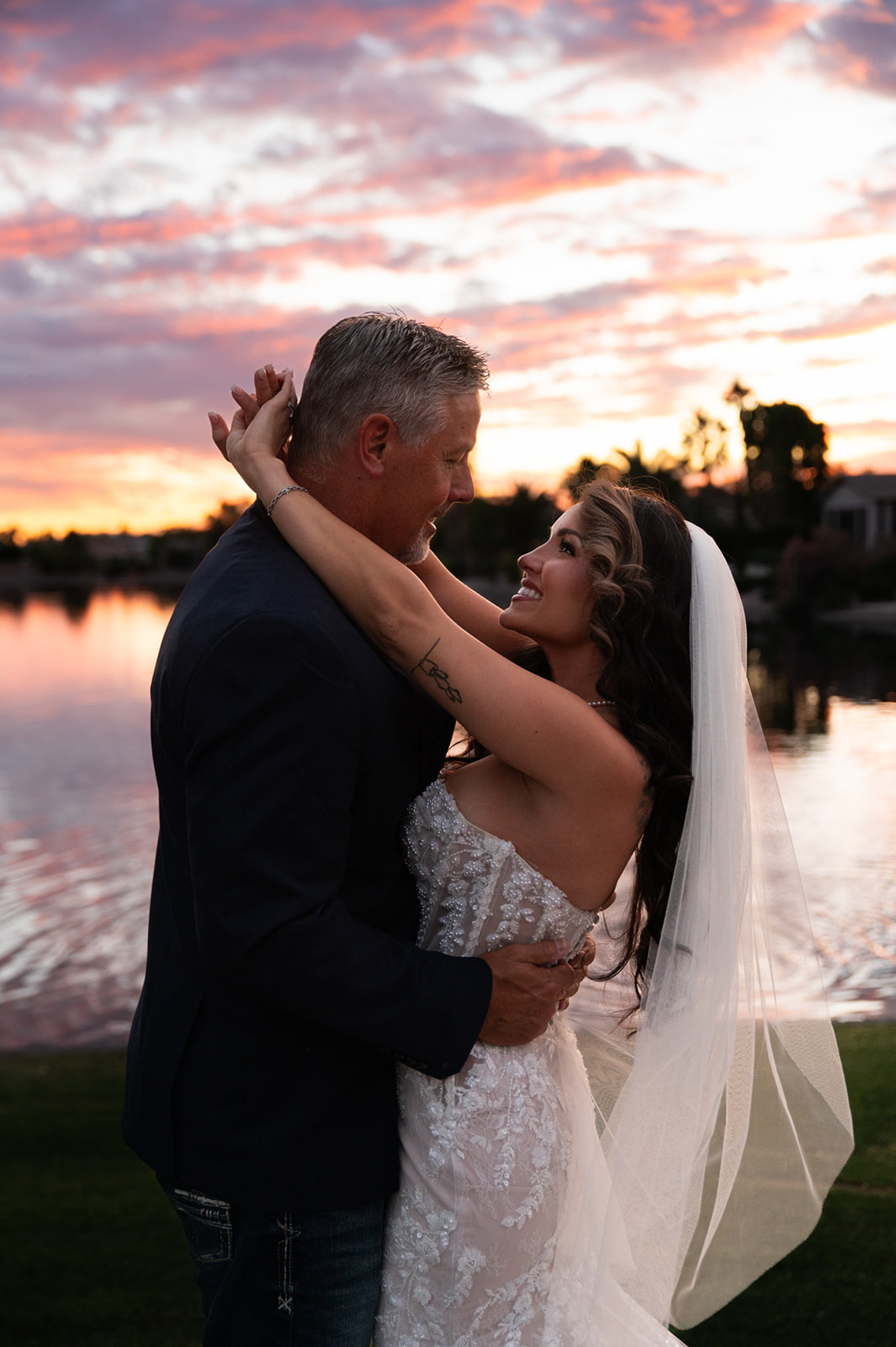 Bride wraps her arms around the groom as they smile at each other beside the water beneath a colorful sunset sky.
