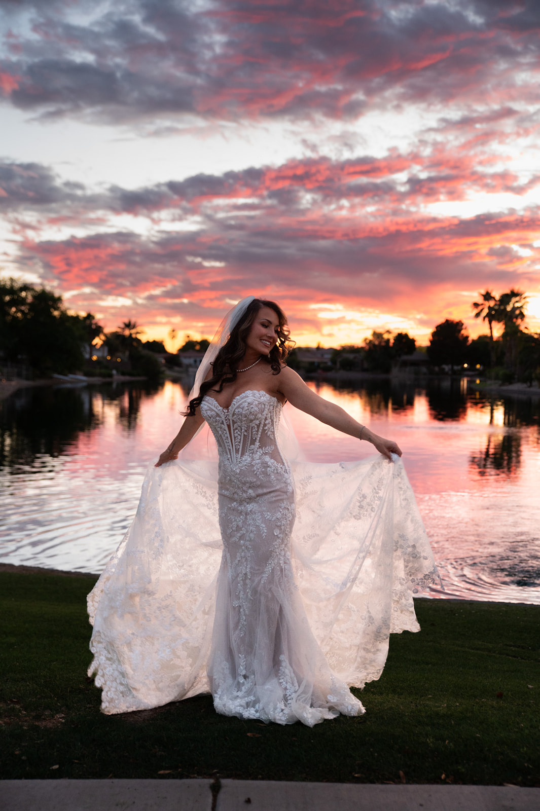 Bride displays her lace wedding gown and veil beside the water at sunset, offering elegant backyard wedding ideas for bridal portraits.
