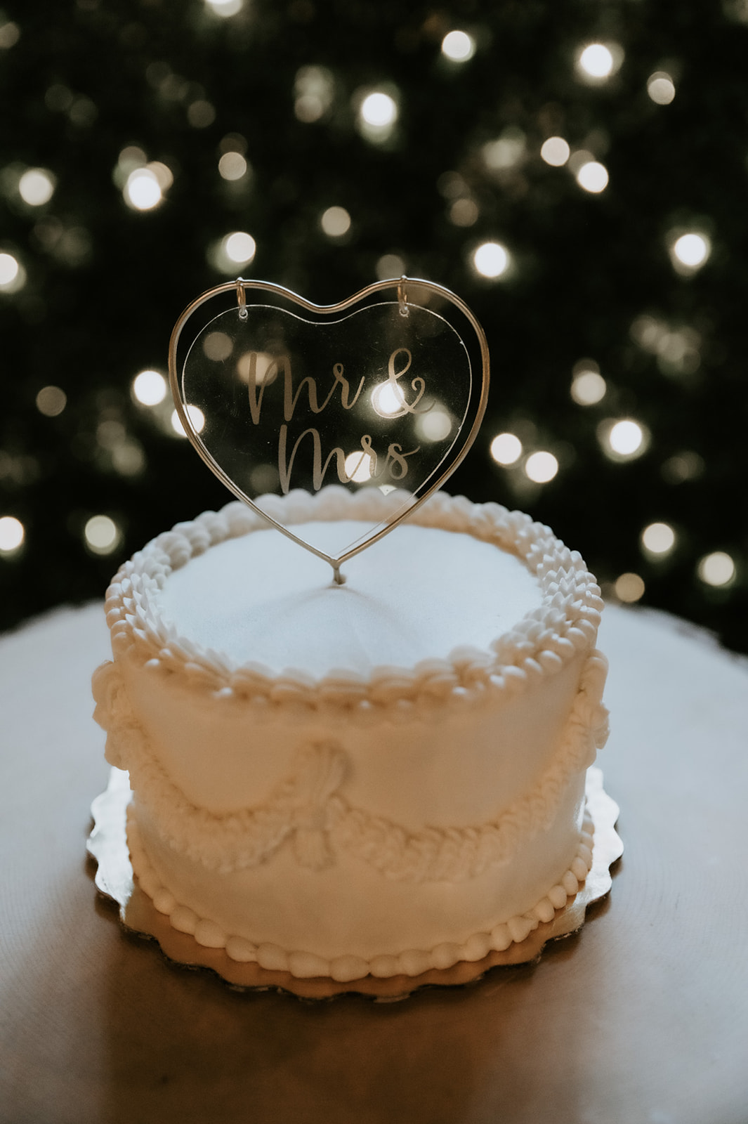 Small white wedding cake with vintage piping and a heart-shaped “Mr & Mrs” topper displayed beneath glowing lights.
