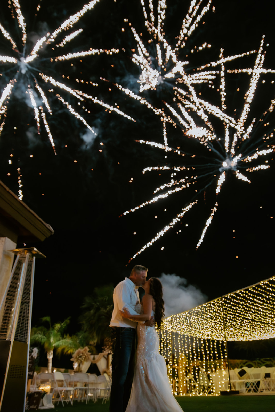 Newlyweds kiss beneath bright fireworks with a glowing canopy of string lights illuminating their outdoor reception.
