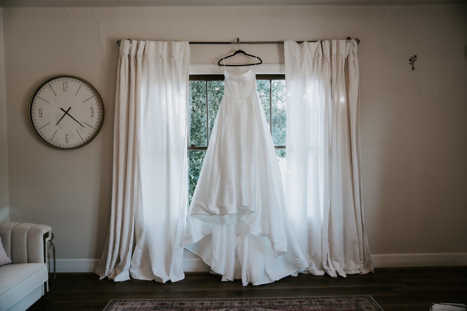White satin wedding dress hangs in front of a bright window before the ceremony.
