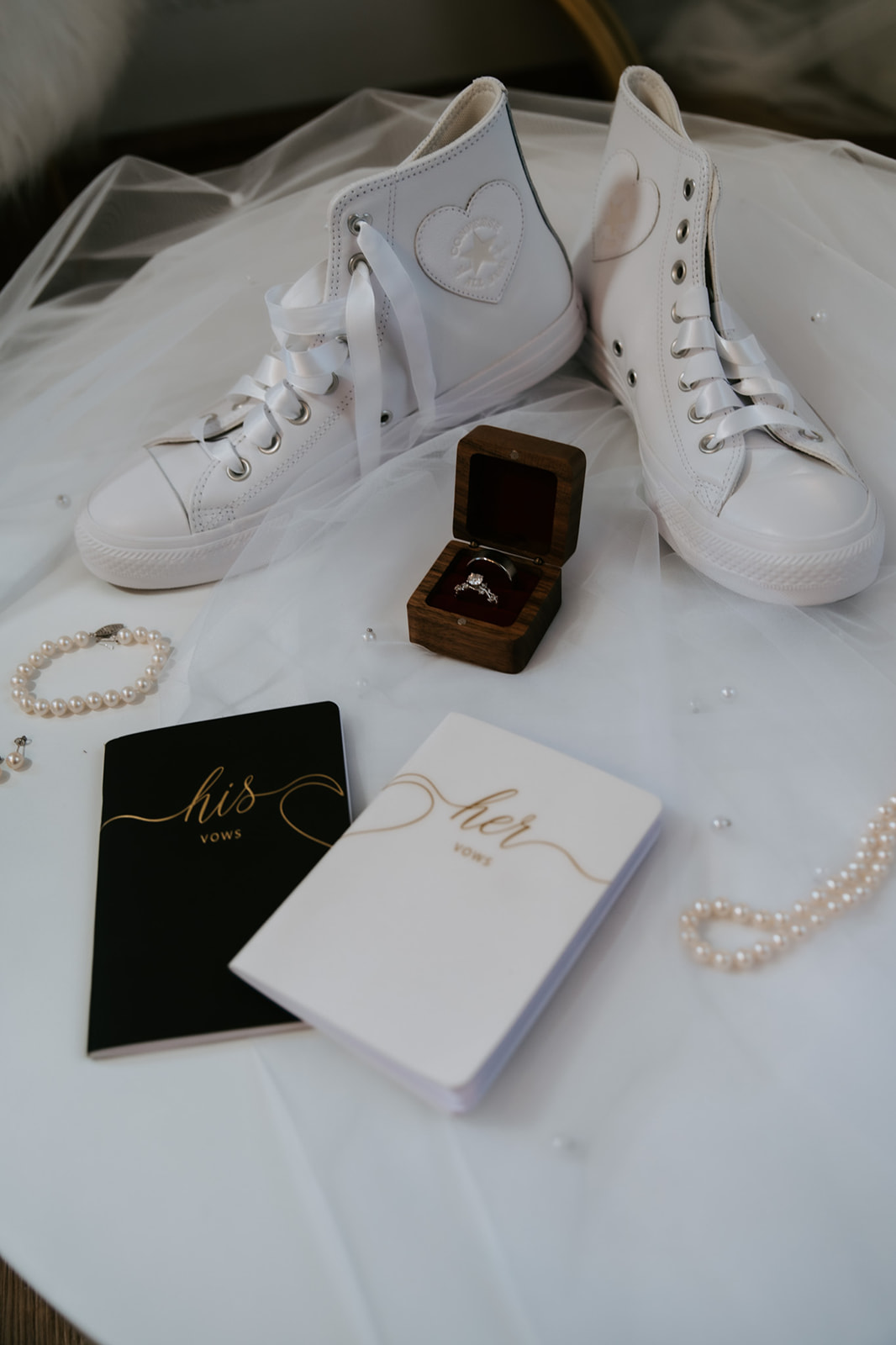 Wedding detail flat lay featuring white high-top sneakers, rings, pearl jewelry, and “His” and “Her” vow books.
