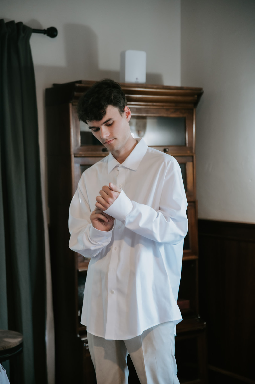 Groom buttons the cuff of his white dress shirt while getting ready.
