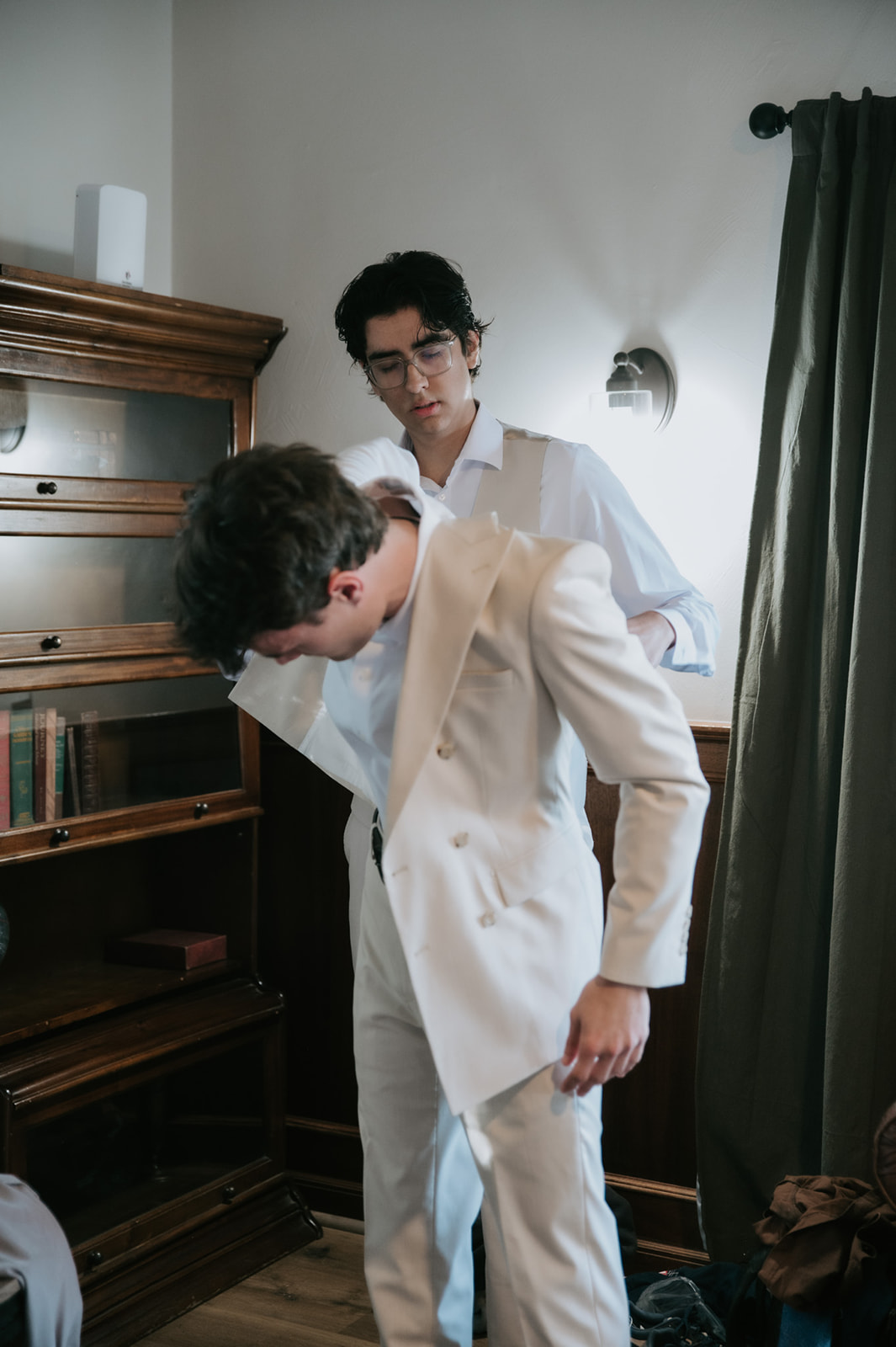Groom gets help putting on his cream double-breasted wedding suit.
