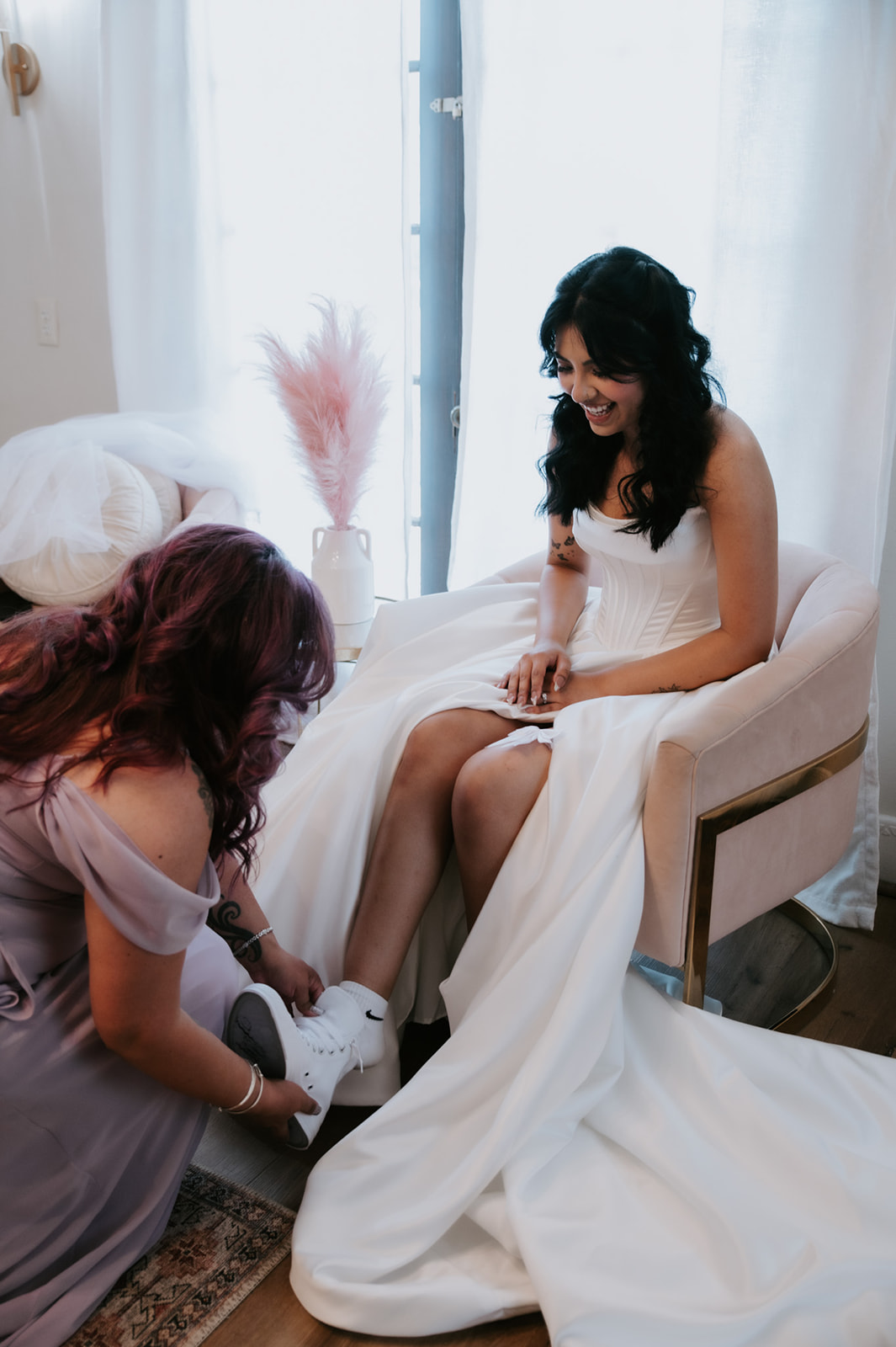 Bride laughs while putting on white sneakers beneath her wedding gown.
