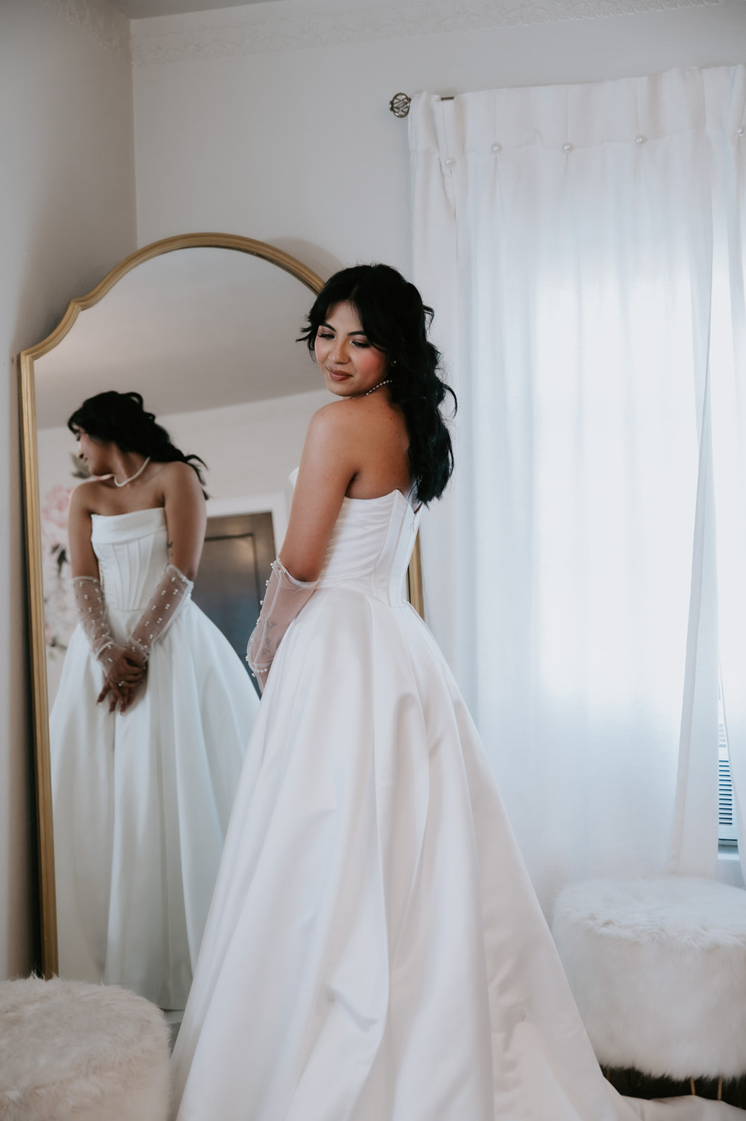 Bride poses beside a full-length mirror in her satin wedding gown and pearl-detailed gloves.
