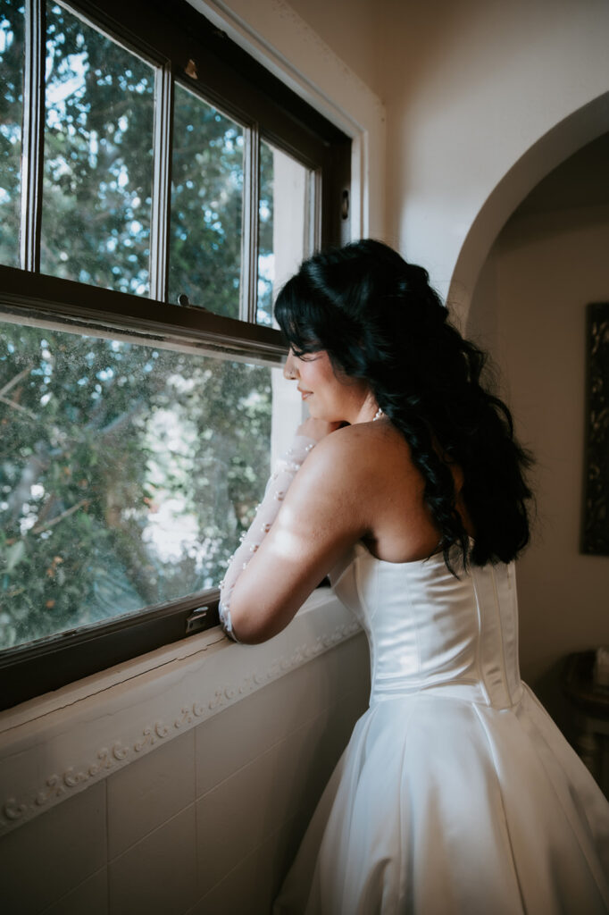Bride looks out a window before her wedding ceremony in a quiet getting-ready moment.
