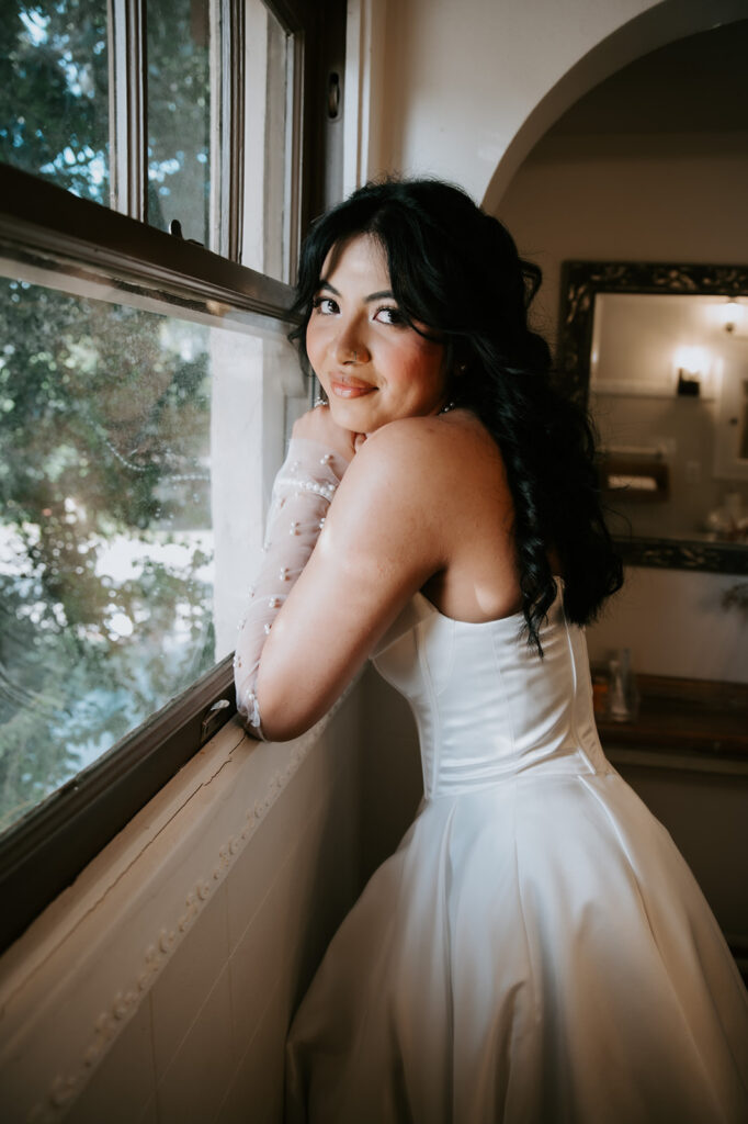 Bridal portrait by a window showcasing the bride’s satin gown, curled hairstyle, and pearl gloves.
