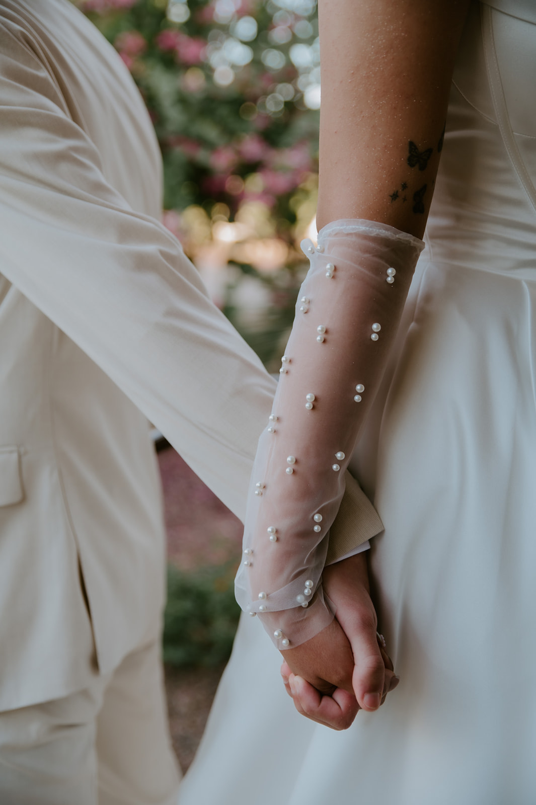 Close-up of the bride and groom holding hands, highlighting the bride’s pearl gloves and butterfly tattoos.
