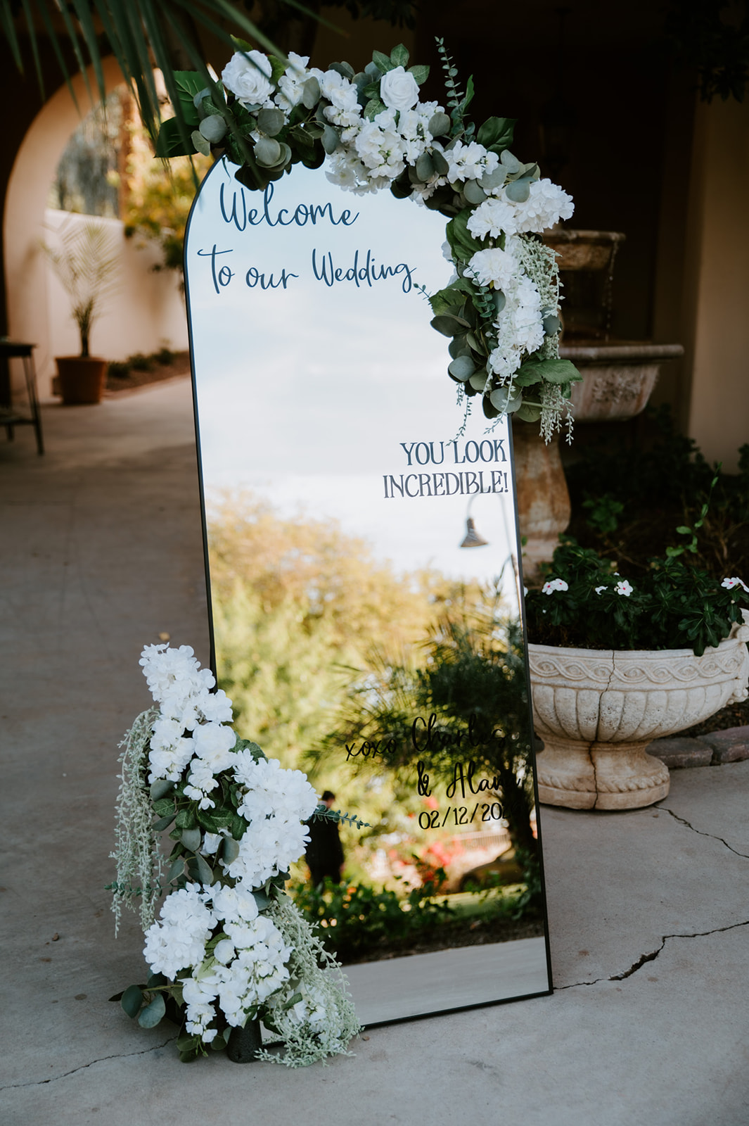 Floral-decorated mirror welcome sign greets guests at an elegant garden wedding.
