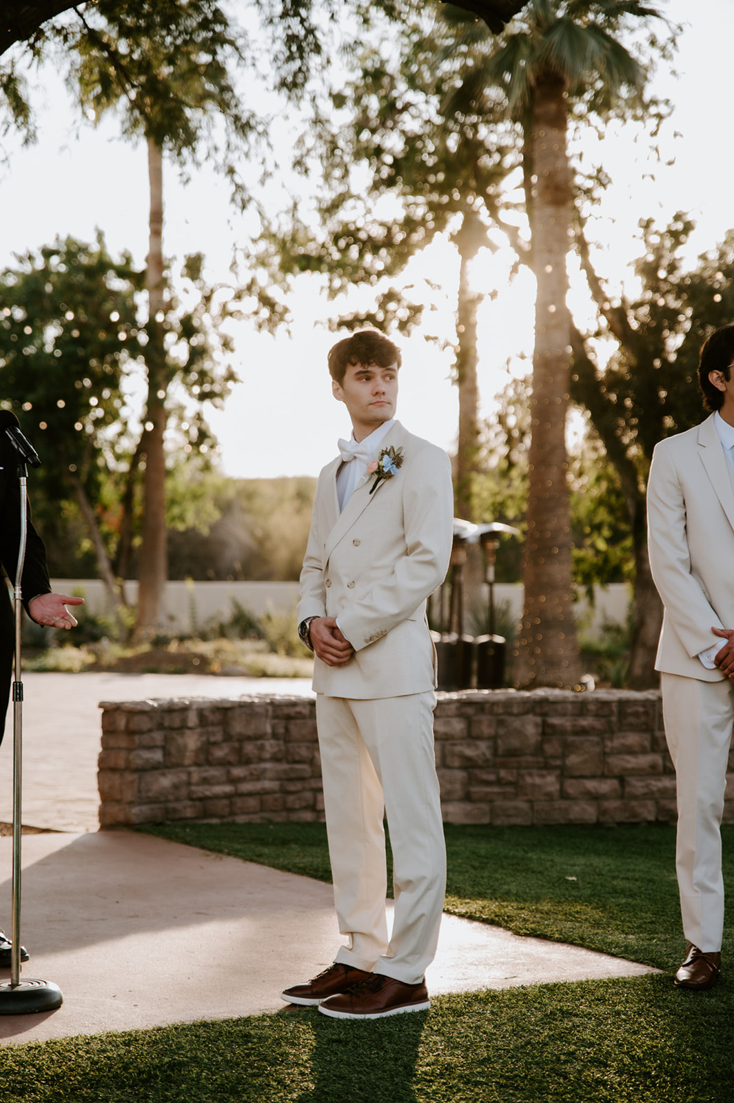 Groom waits at the ceremony altar in a cream suit and bow tie during golden hour.
