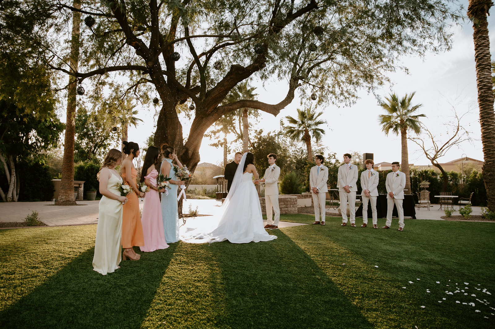 Wedding party surrounds the bride and groom beneath a large tree during their outdoor ceremony at Secret Garden by Wedgewood Weddings.
