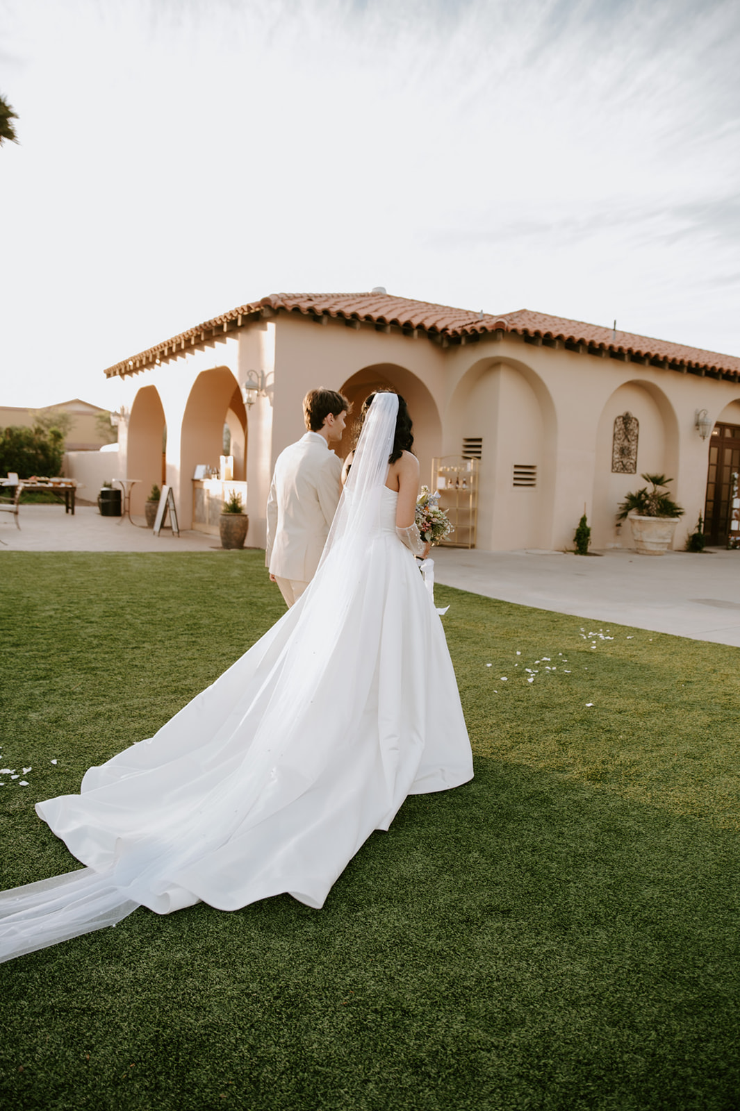 Bride and groom walk together across the lawn, showcasing the bride’s long flowing wedding gown and veil.
