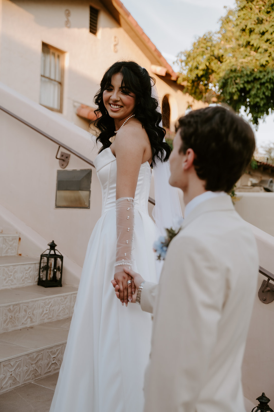 Bride smiles back at the groom while holding his hand on an outdoor staircase.
