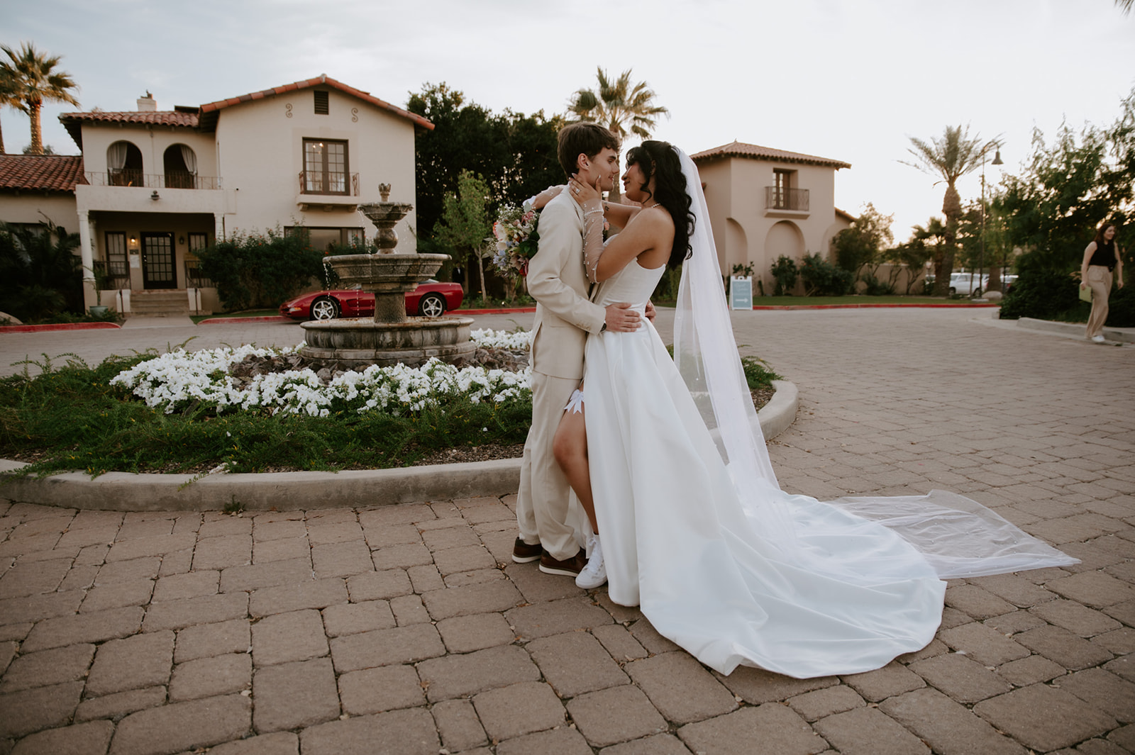 Bride and groom embrace near a courtyard fountain with Mediterranean-style architecture in the background.
