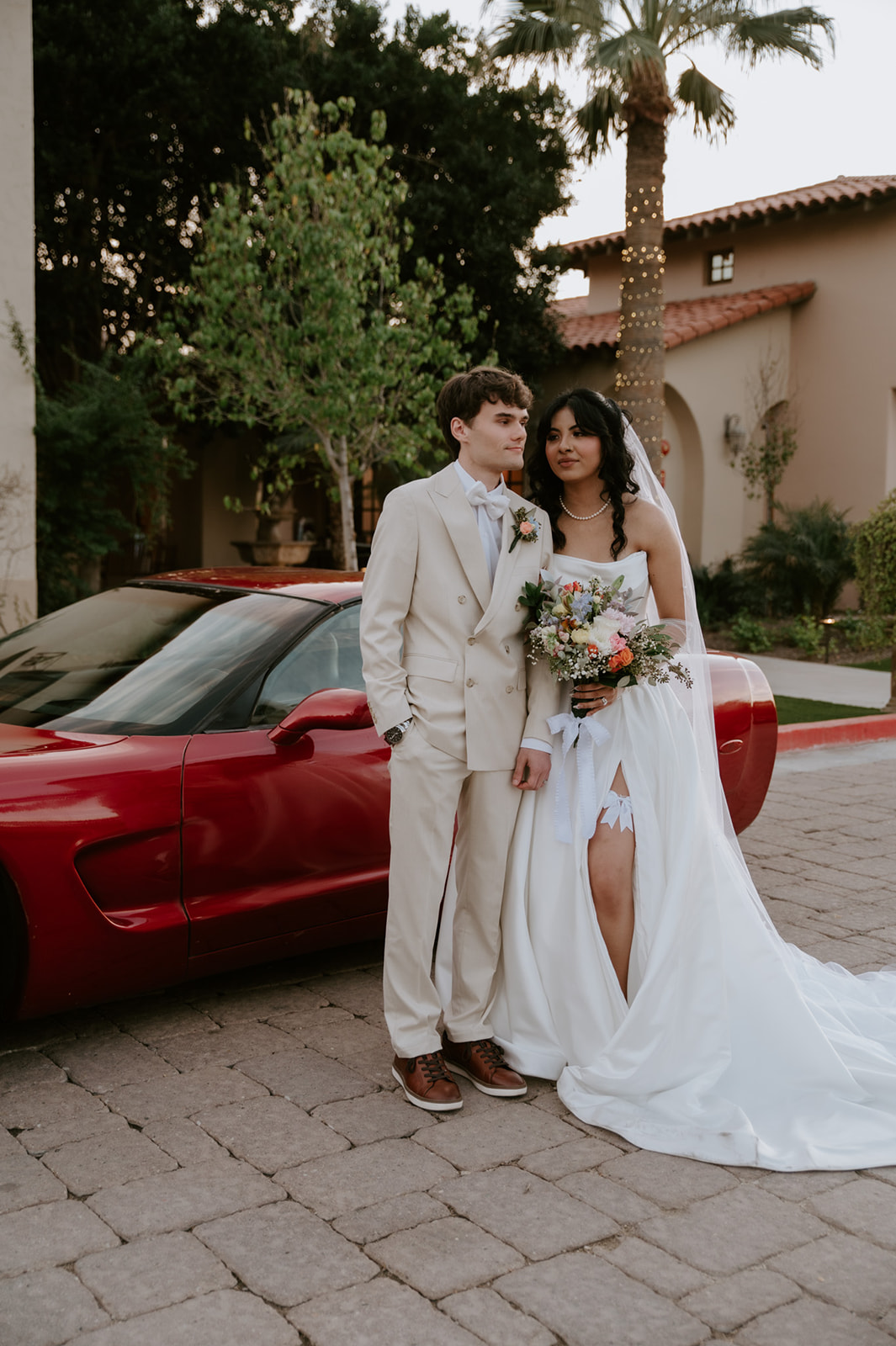 Bride and groom pose with a colorful bouquet beside a red sports car at Secret Garden by Wedgewood Weddings.

