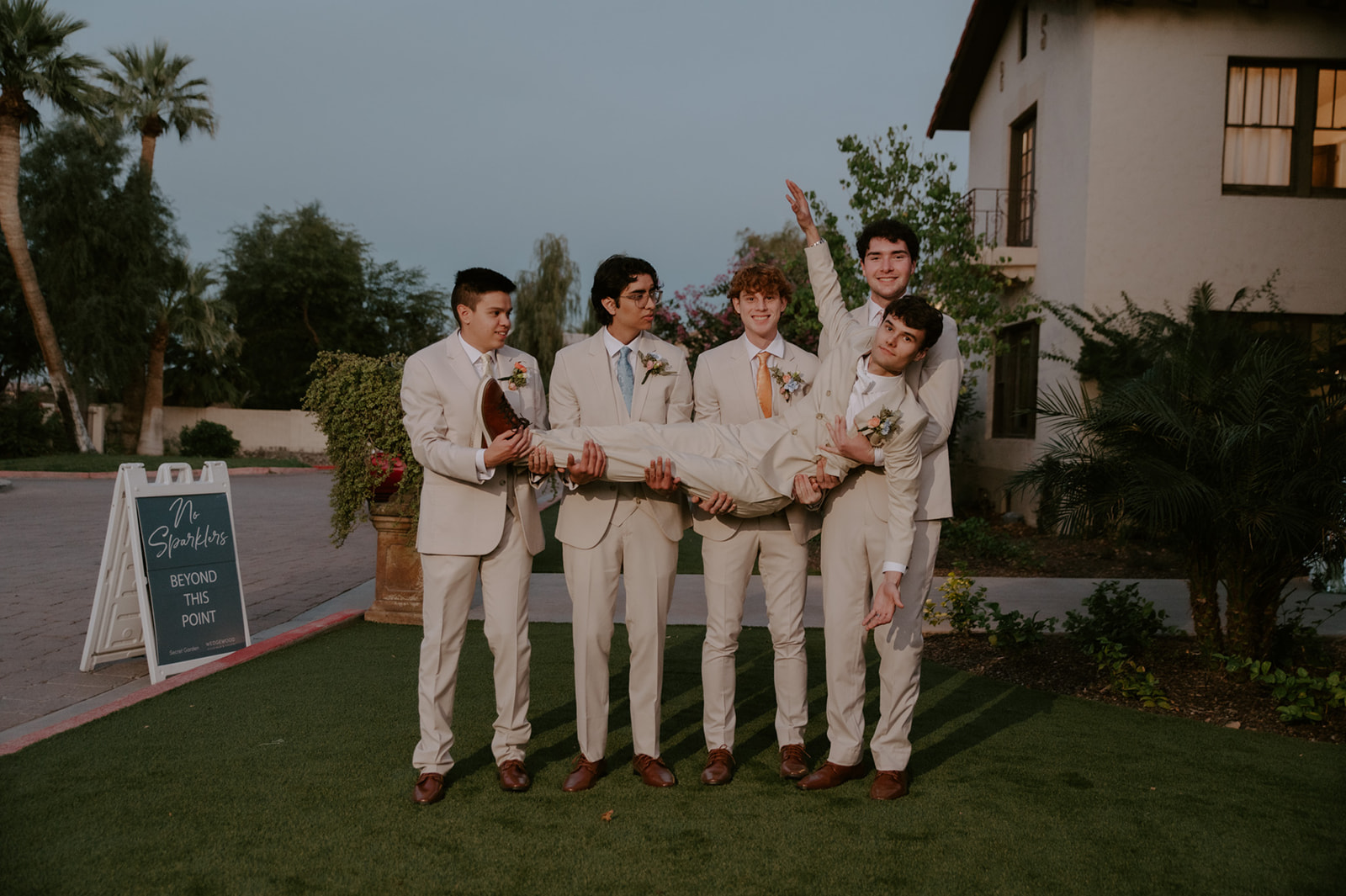 Groom and groomsmen in light neutral suits pose playfully together outdoors.
