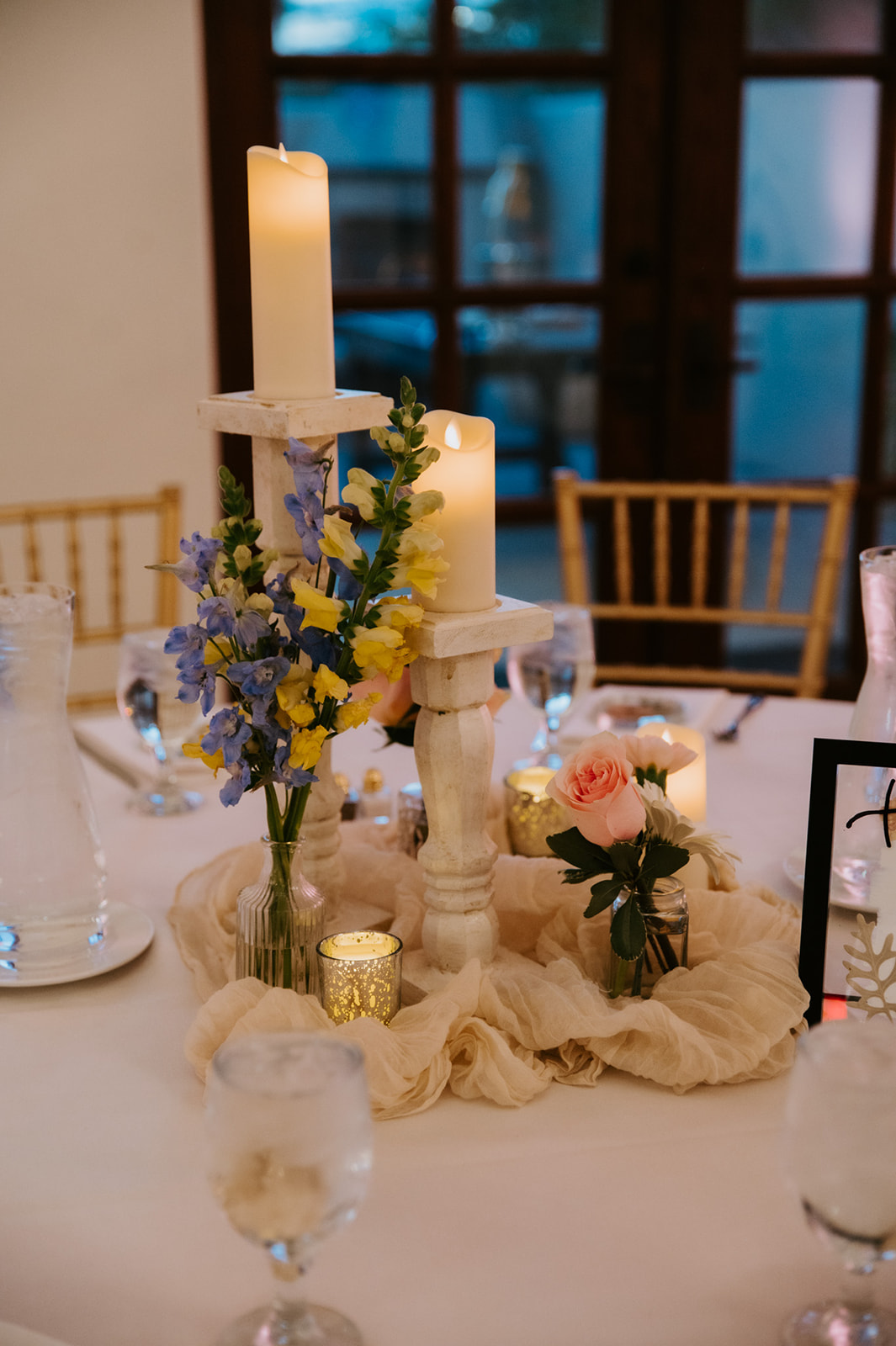 Candlelit wedding centerpiece with blue, yellow, and peach flowers arranged on a reception table.
