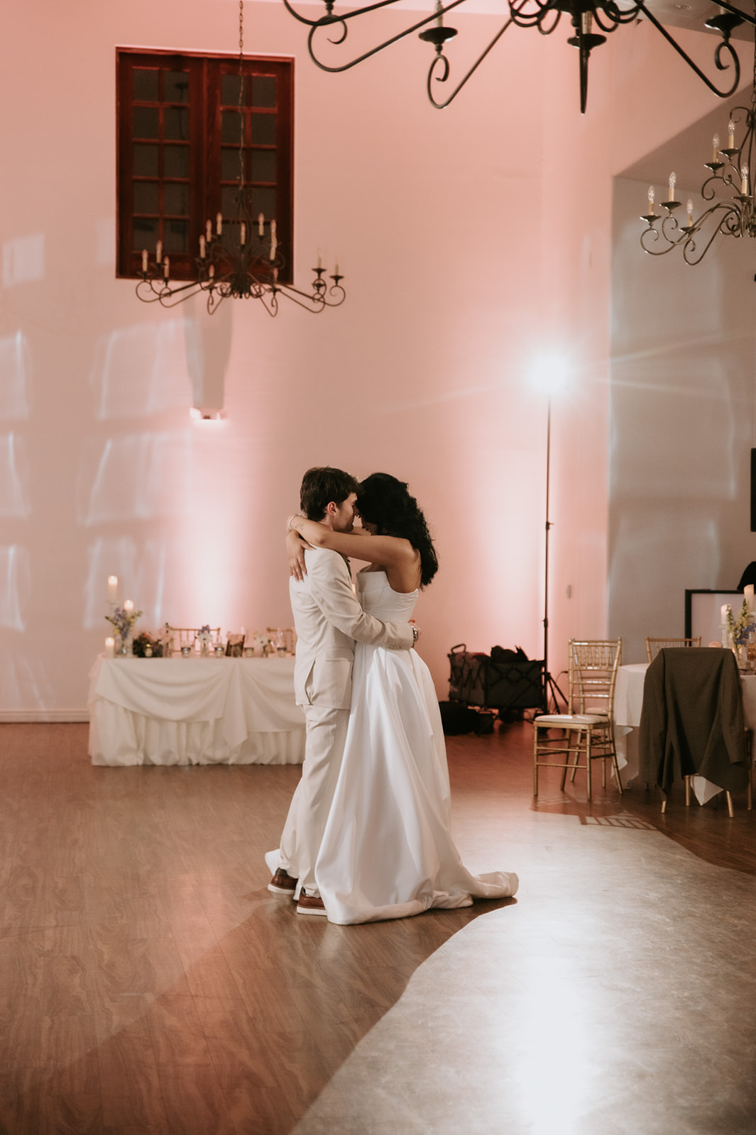 Bride and groom embrace on the dance floor beneath chandeliers during their wedding reception.
