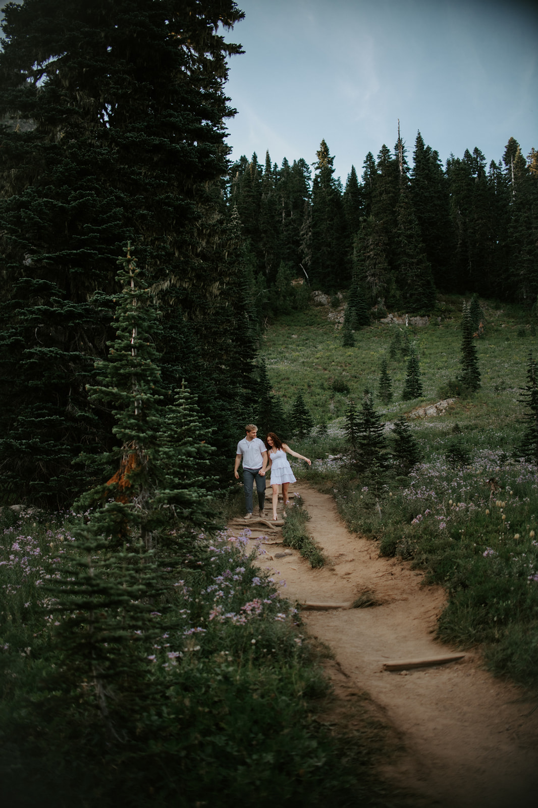 Couple eloping in Washington walking hand in hand along a mountain trail surrounded by wildflowers and evergreen trees.