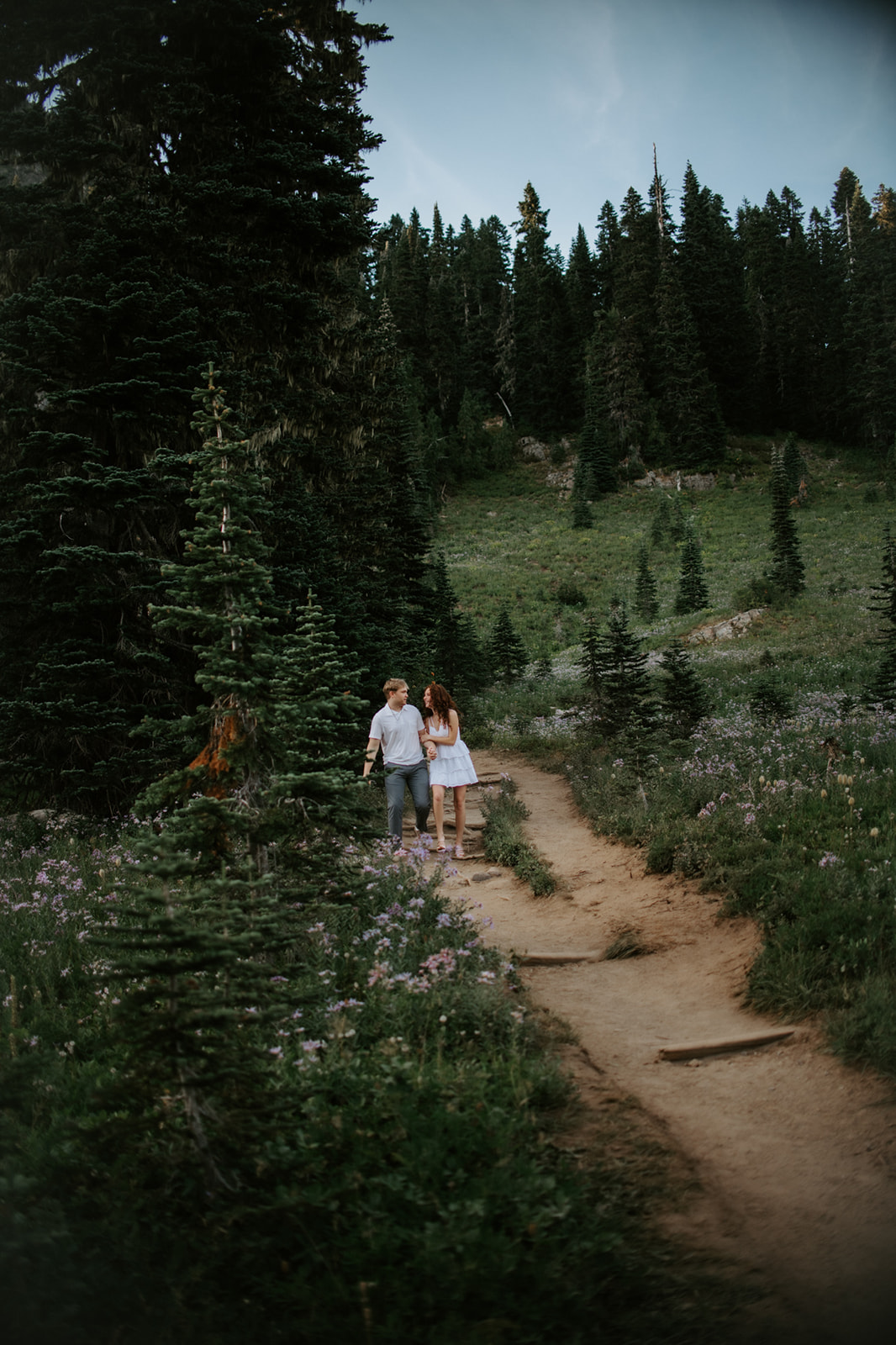 Couple eloping in Washington walking together along a mountain trail surrounded by wildflowers and towering evergreen trees.