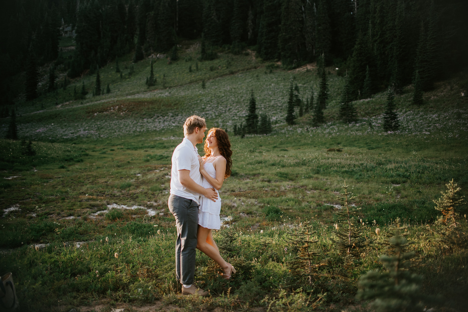 Couple embracing in a wildflower-filled mountain meadow surrounded by evergreen trees during golden hour.