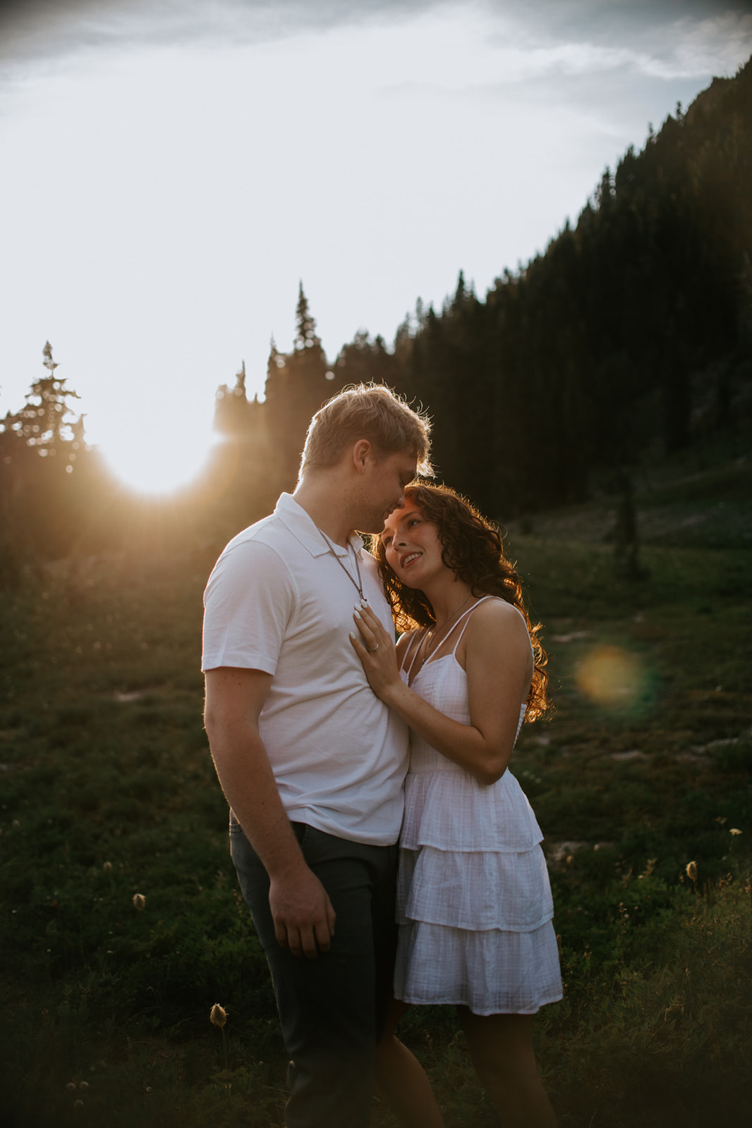 Couple embracing in a mountain meadow during golden hour with evergreen-covered hills in the background.