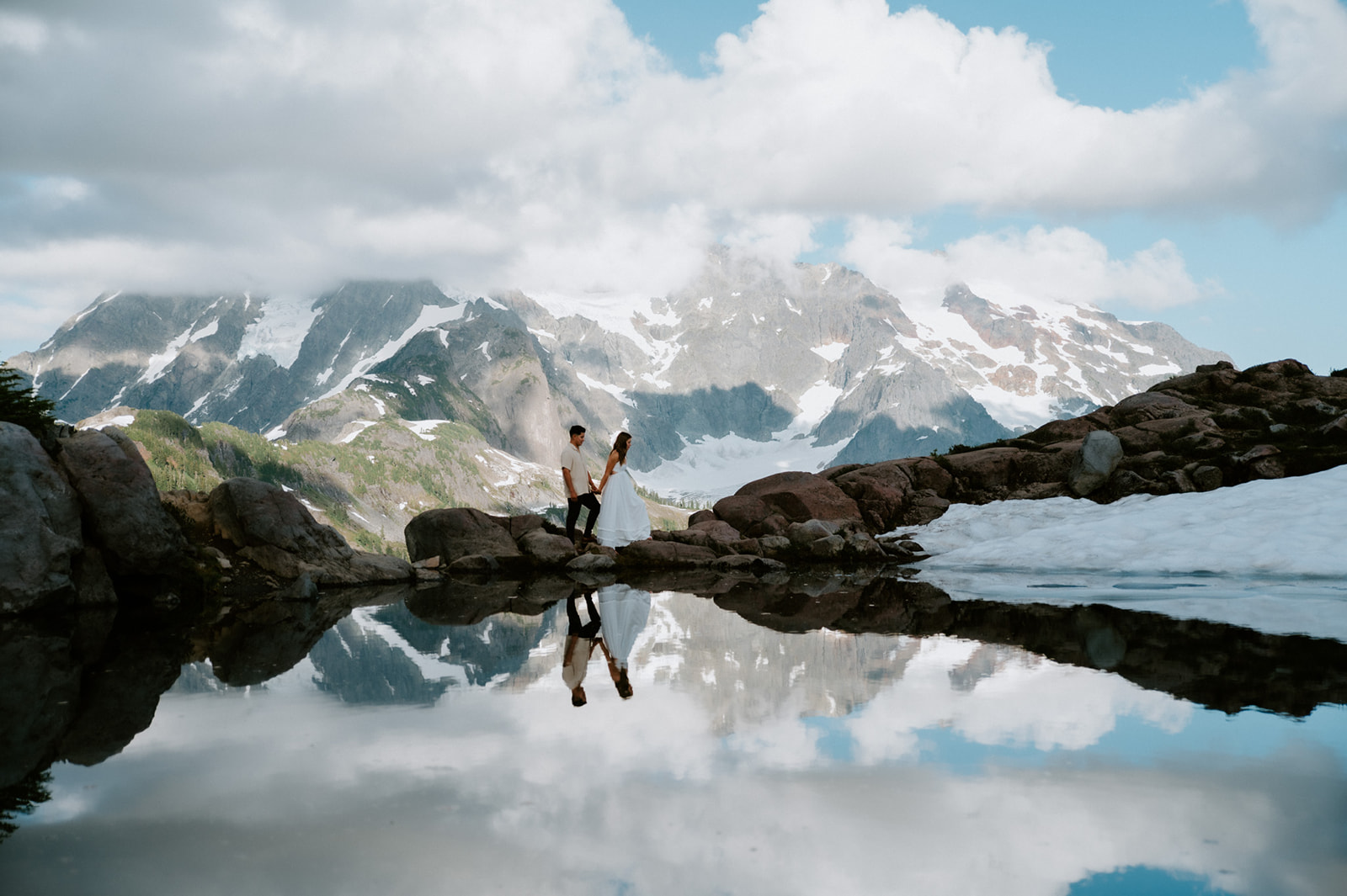 Couple exploring rocky alpine terrain beside a reflective pond with dramatic snow-covered Washington mountains in the background.