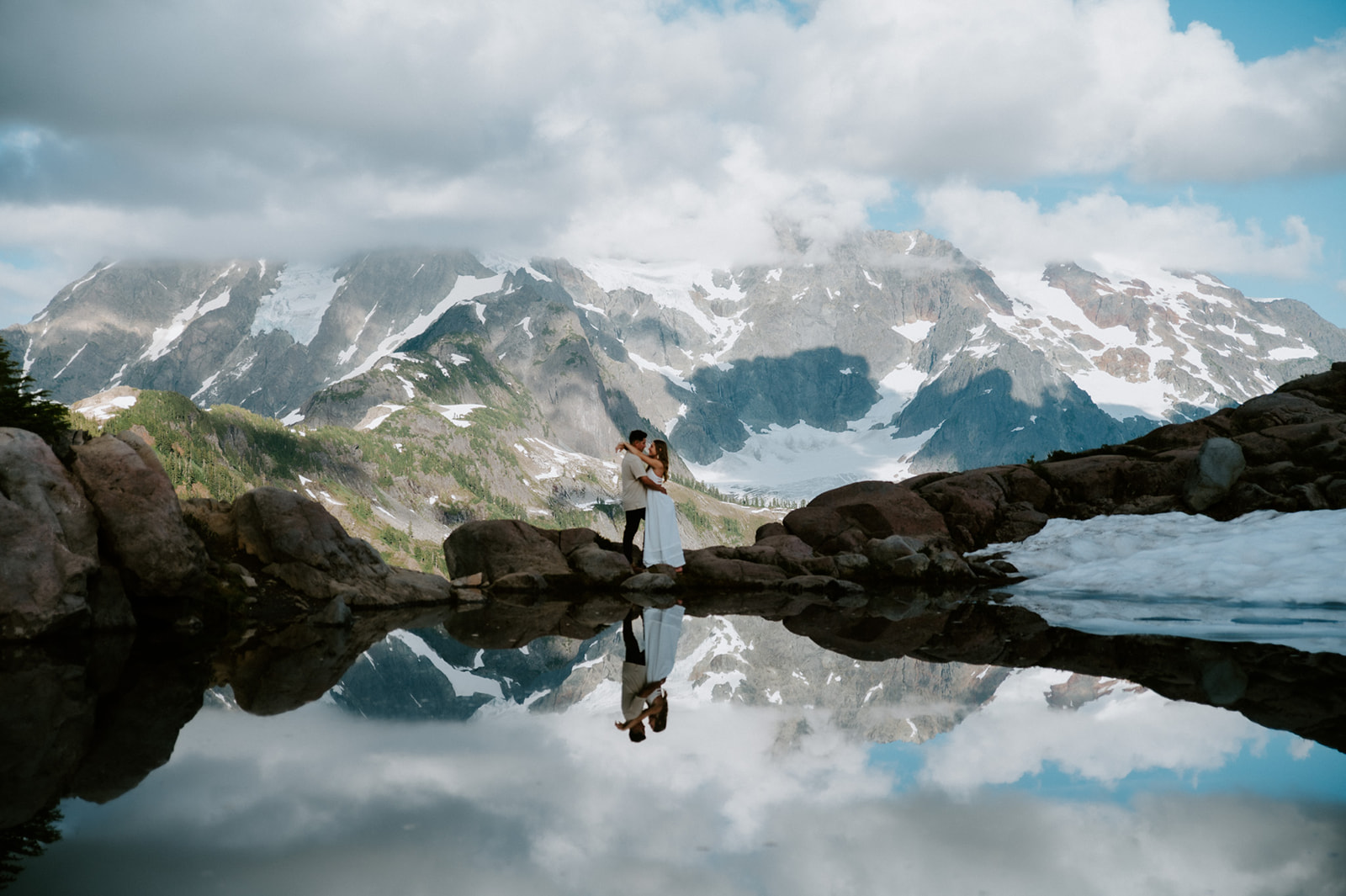Couple eloping in Washington embracing beside an alpine lake with dramatic snowy mountains reflected in the water.