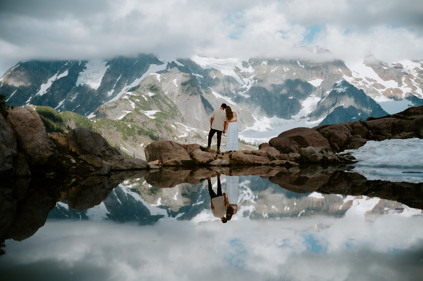 Couple eloping in Washington overlooking a dramatic snow-covered mountain range reflected in a clear alpine lake.