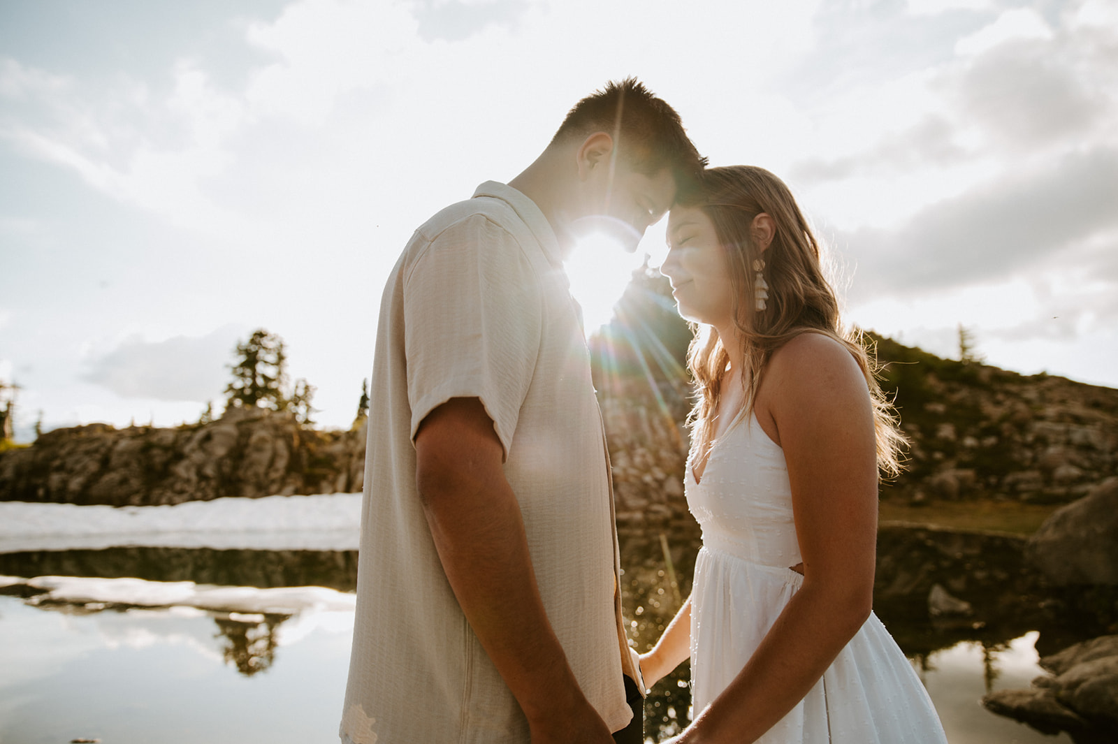 Couple standing forehead to forehead as golden sunlight shines between them beside a reflective mountain pond.