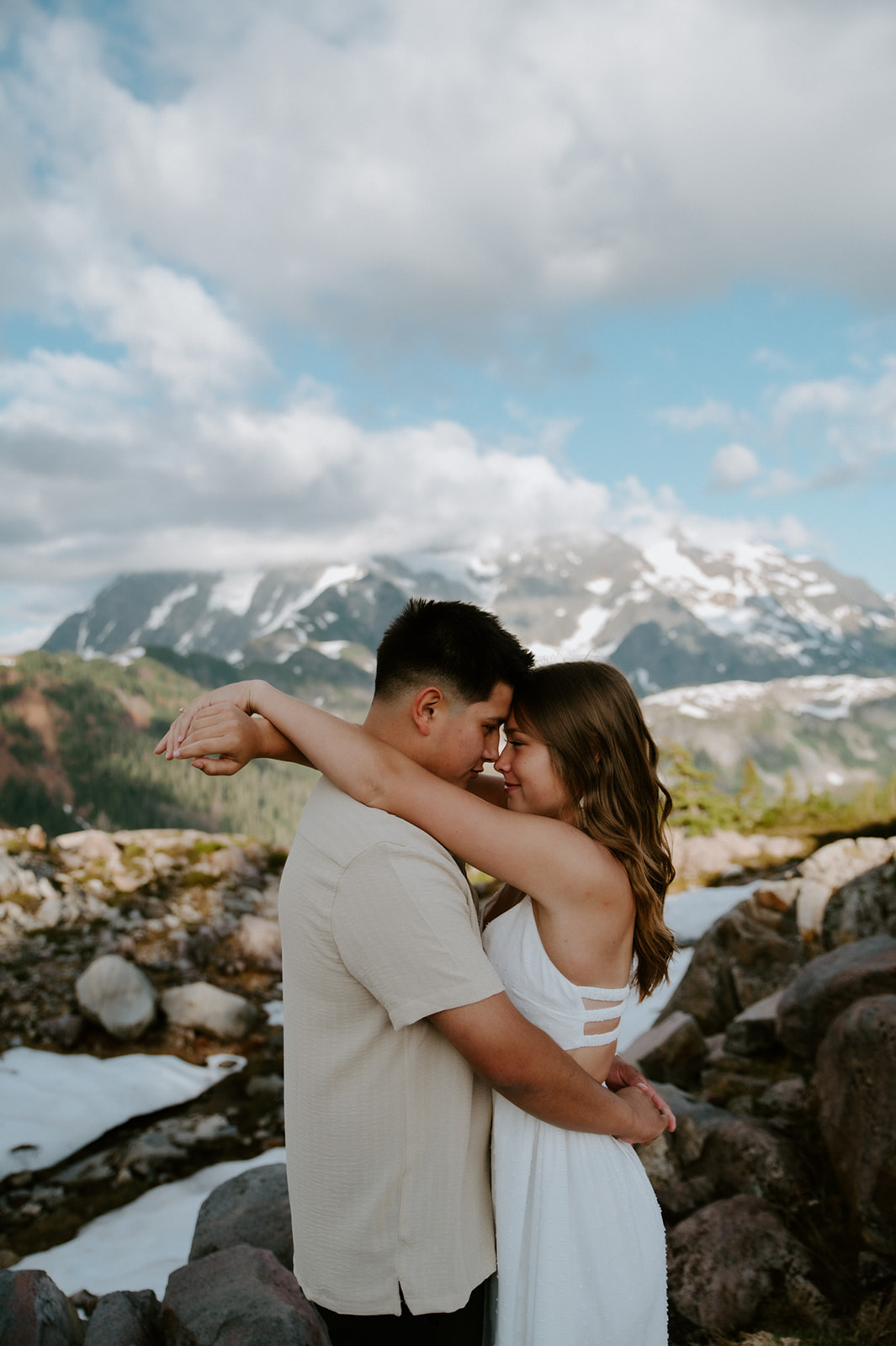 Couple embracing on a rocky mountain trail with patches of snow and dramatic peaks in the background.