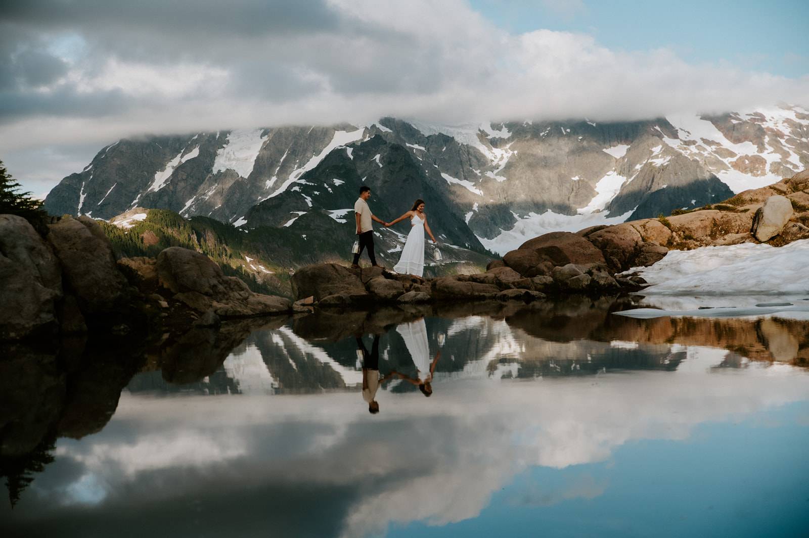 Couple holding hands beside a reflective alpine pond with dramatic snow-covered mountains and clouds in the background.