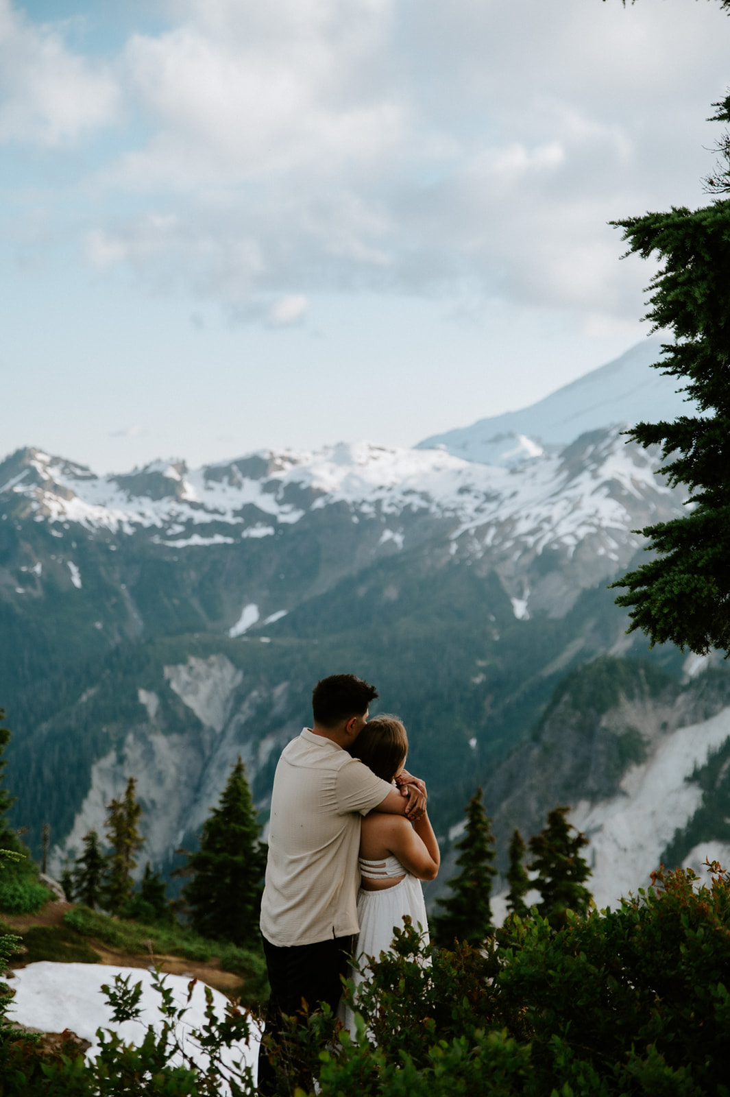 Couple embracing while looking out at dramatic snow-covered mountain peaks surrounded by evergreen trees.