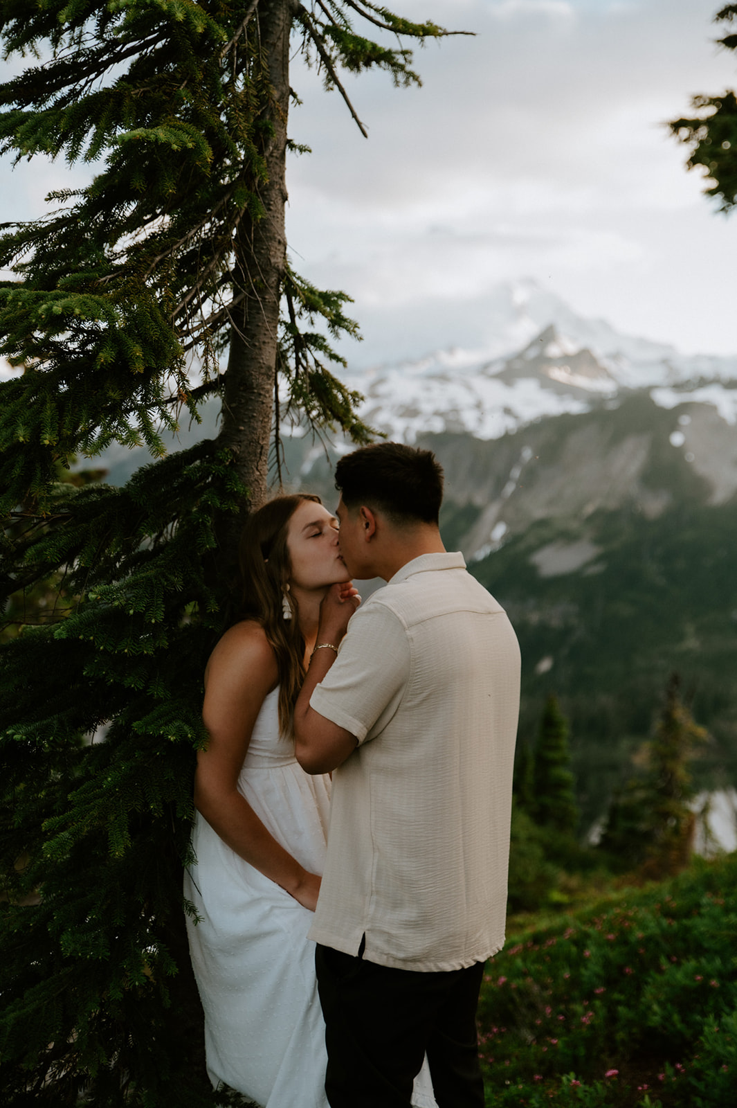 Couple kissing beneath an evergreen tree with dramatic snow-covered Washington mountains in the background.