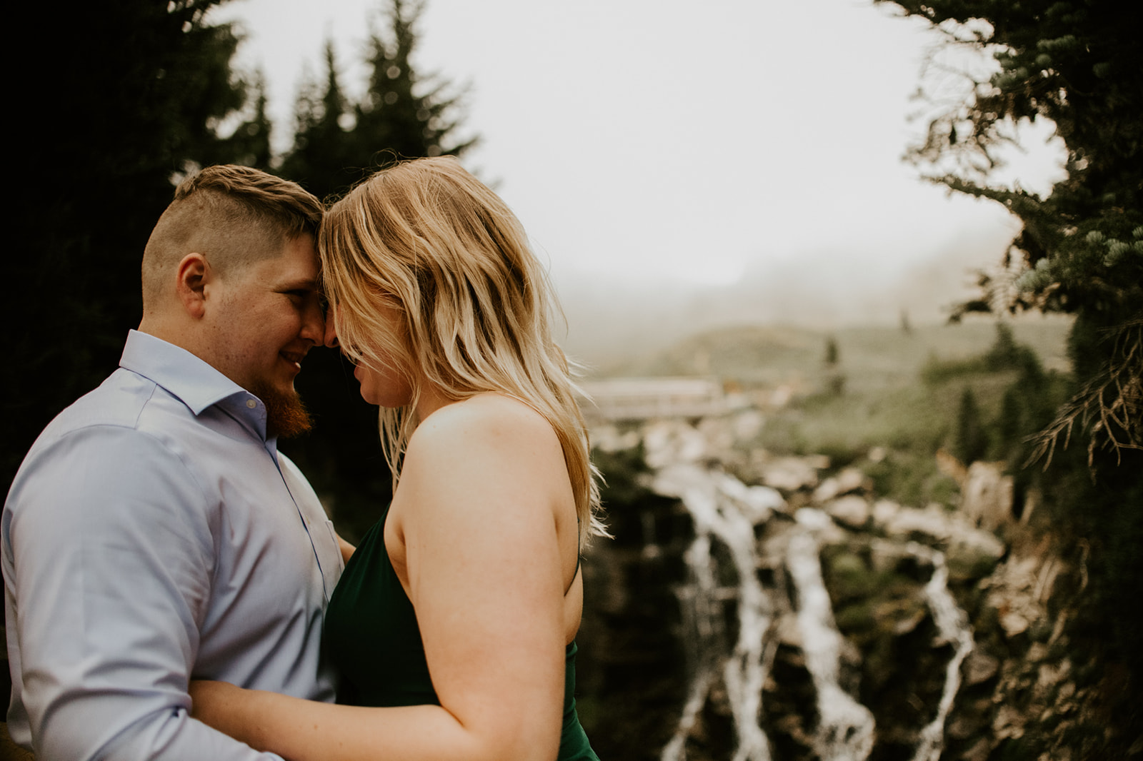 Couple embracing beside a cascading waterfall surrounded by evergreen trees and misty mountain scenery.