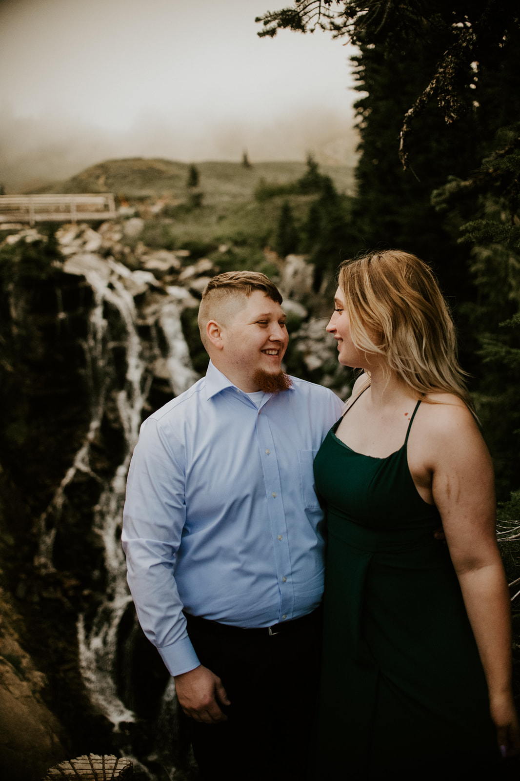 Couple eloping in Washington smiling together with a cascading waterfall and moody alpine landscape behind them.