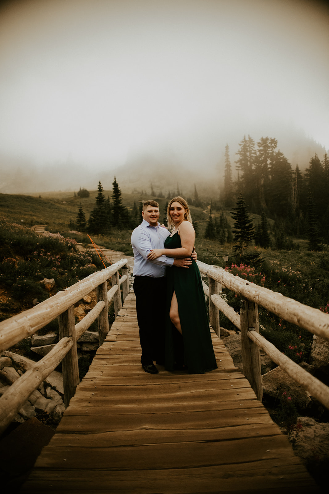 Couple posing together on a wooden mountain bridge surrounded by fog, wildflowers, and evergreen trees.